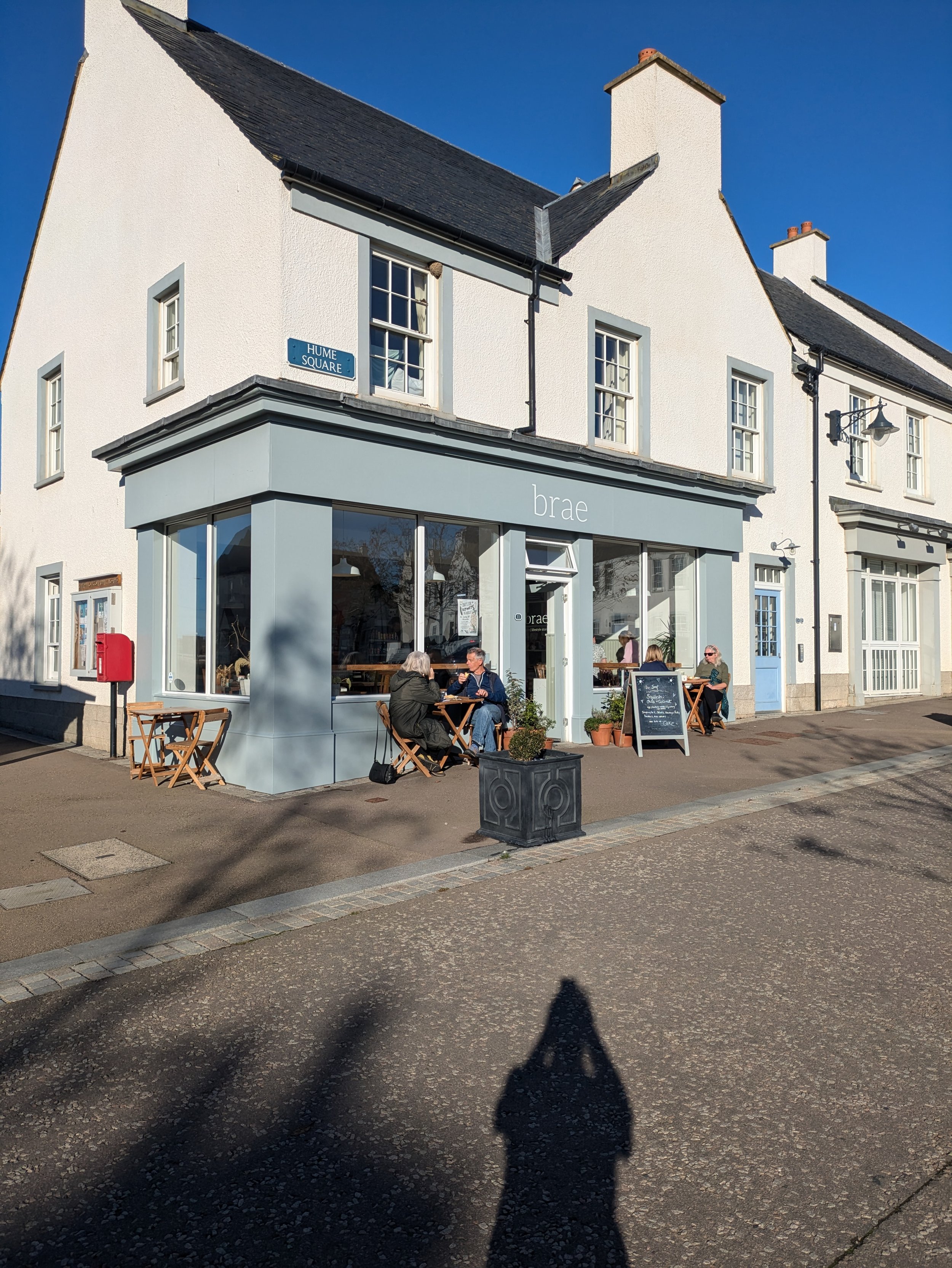 Exterior of a cafe named 'brae' with outdoor seating on a sunny day, white building with large windows and light blue accents, people sitting at tables, a chalkboard menu, and a street sign that says 'Hume Square'.  Takeaway menu.