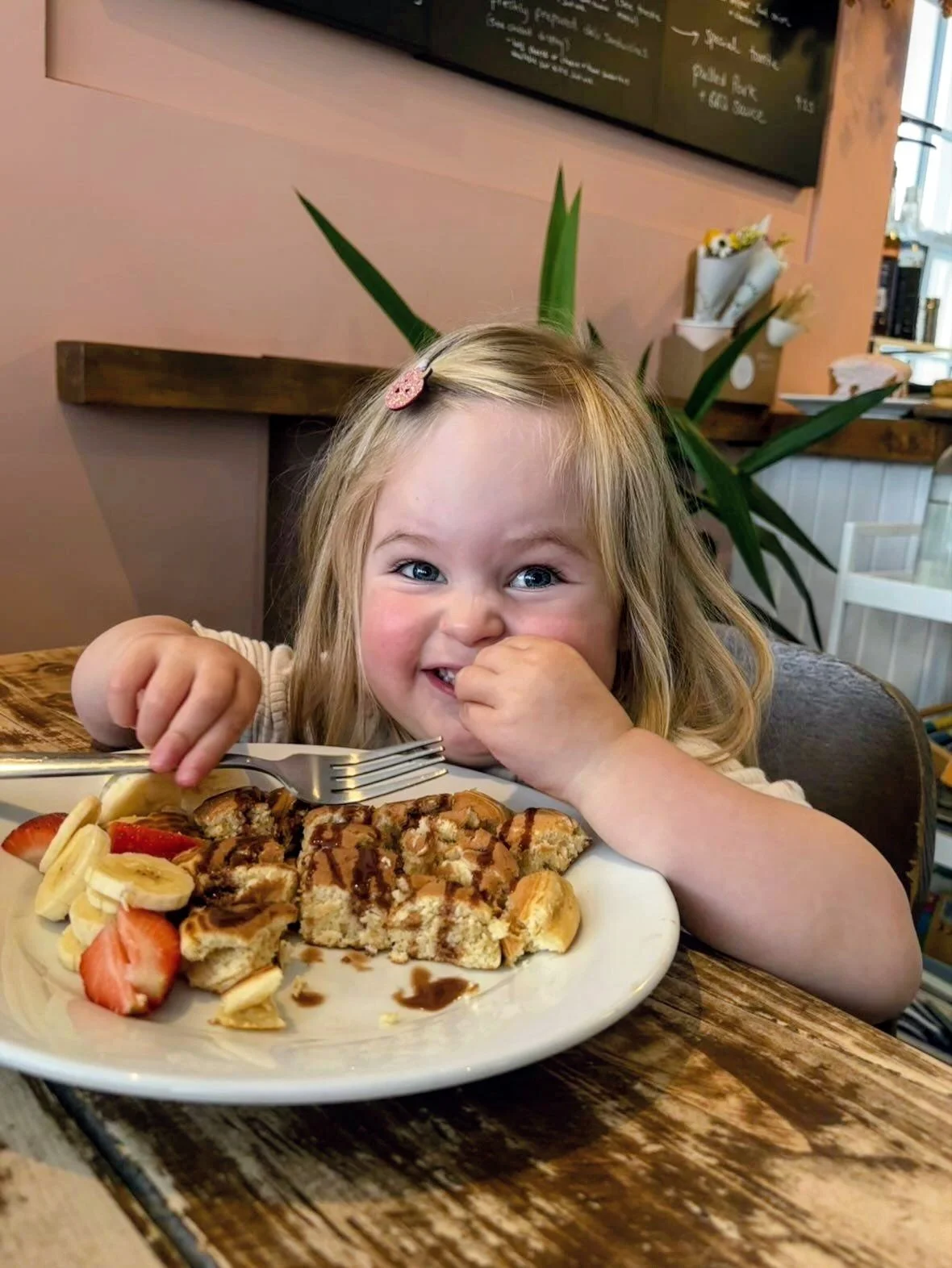 A young girl with blonde hair and a pink hair clip, smiling and eating a piece of cake with strawberries, banana slices, and chocolate drizzle at a restaurant or cafe.