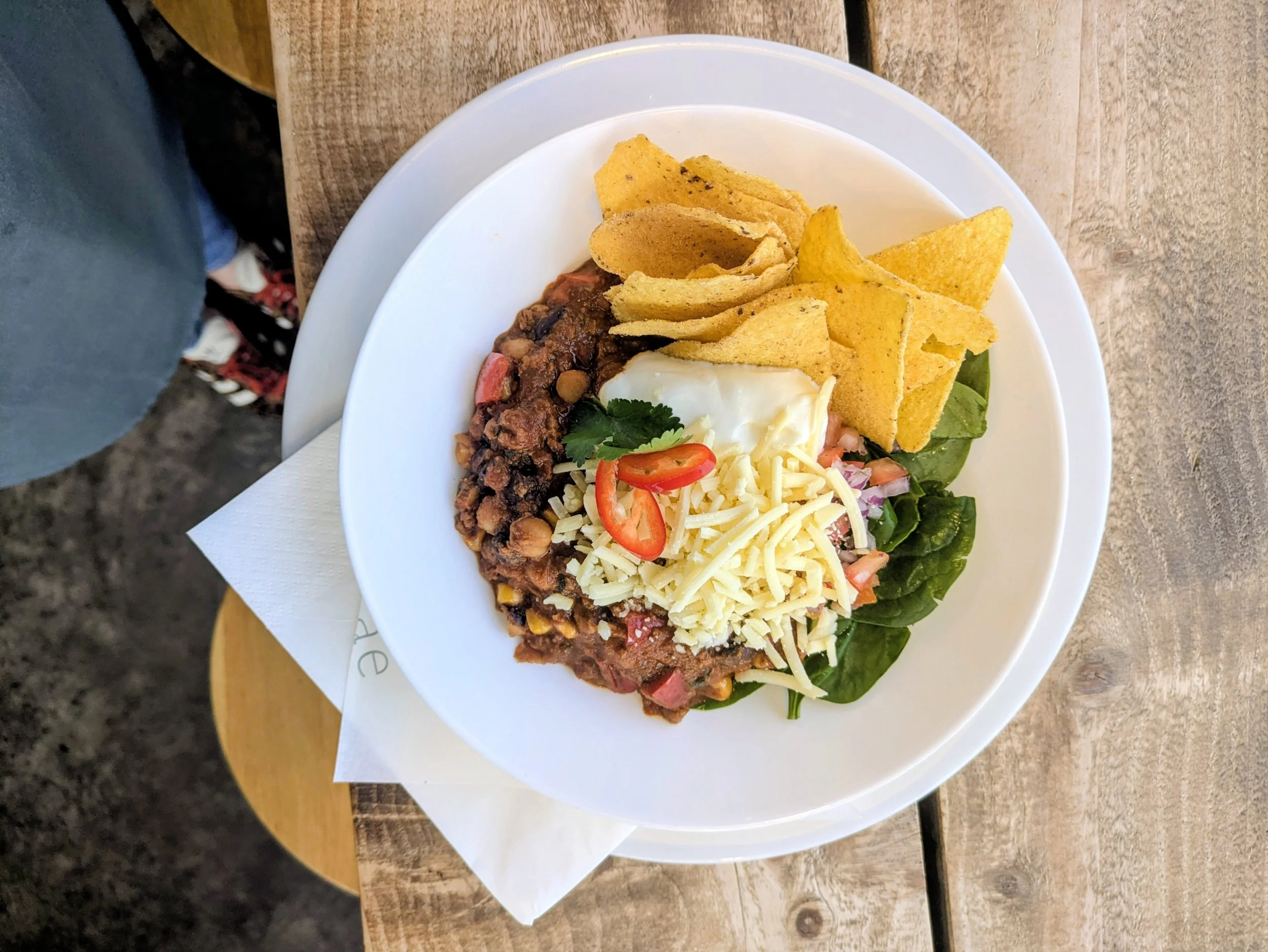 Bowl of chili with shredded cheese, diced tomatoes, and chili peppers, served with tortilla chips, a poached egg, and fresh spinach on the side.