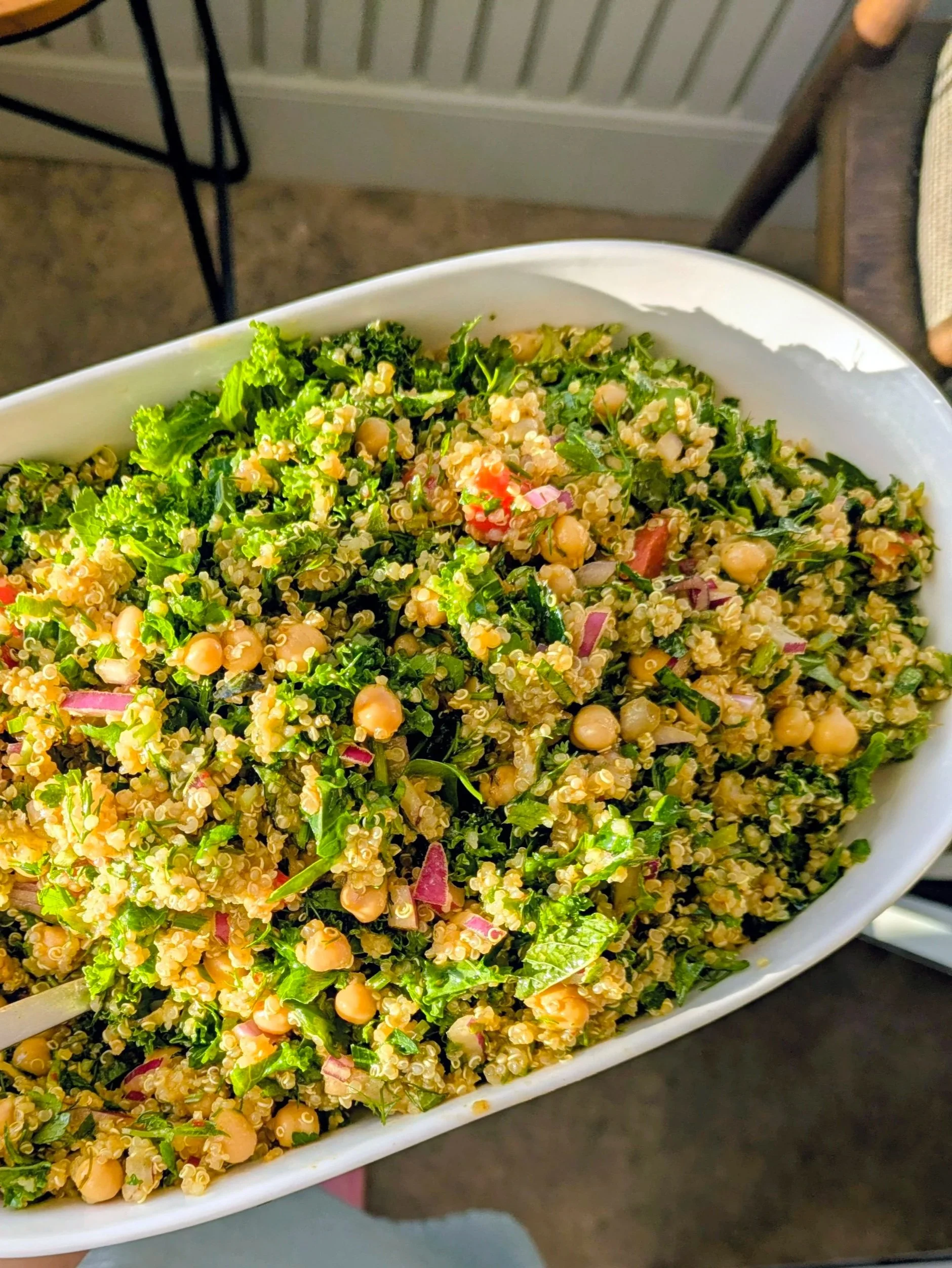 A large white dish filled with a colorful salad containing kale, quinoa, chickpeas, red onion, tomatoes, and herbs.