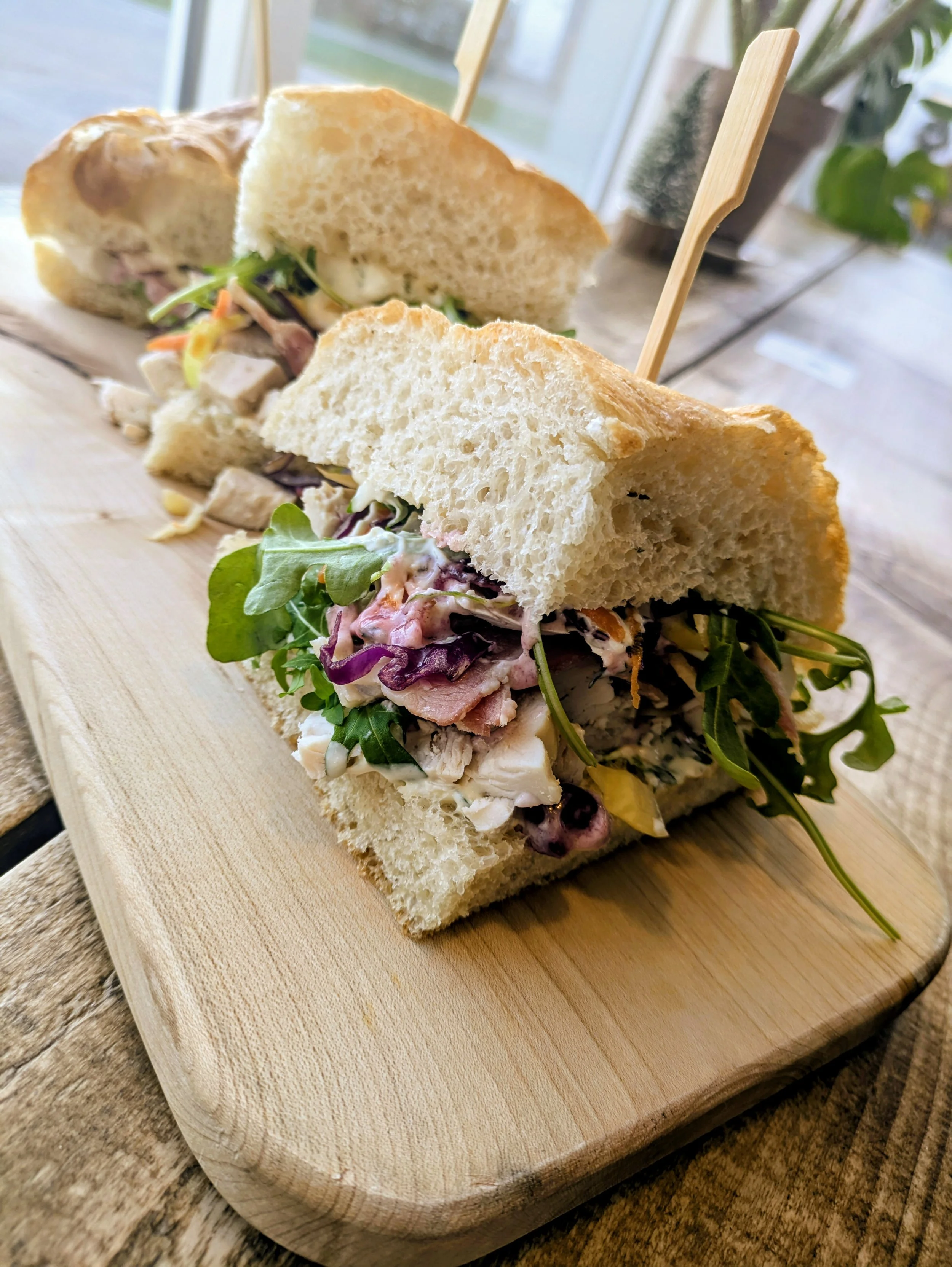 Close-up of a sandwich on a wooden cutting board, with fresh bread, turkey, bacon, lettuce, and creamy dressing, set on a rustic table near a window.