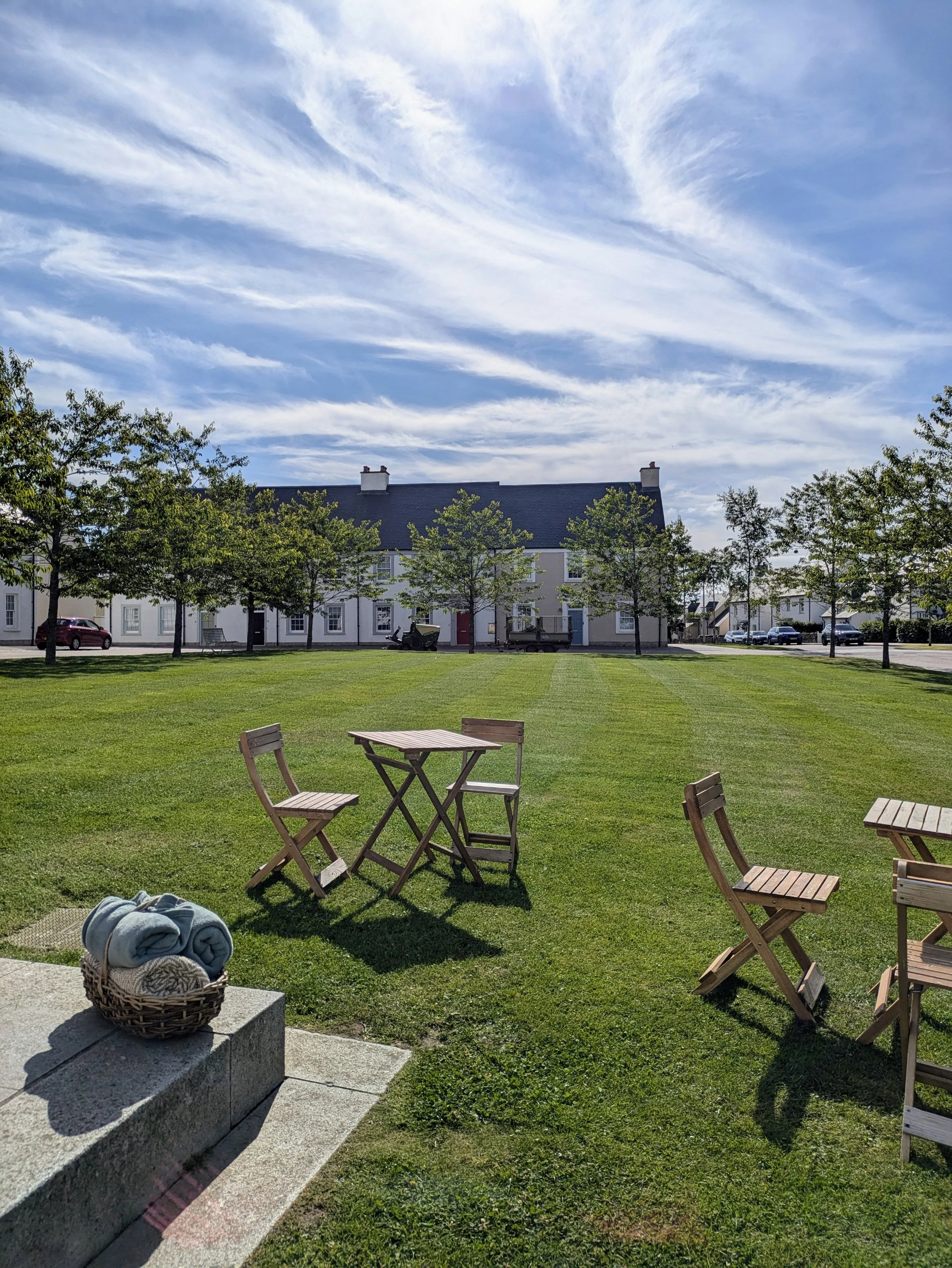 Outdoor scene with a grassy lawn, wooden chairs and tables, a basket of rolled towels, trees, residential buildings, and a partly cloudy blue sky.  Hume Square Chapelton surrounded by free parking and close to cafe.  Disabled parking, phone box, post