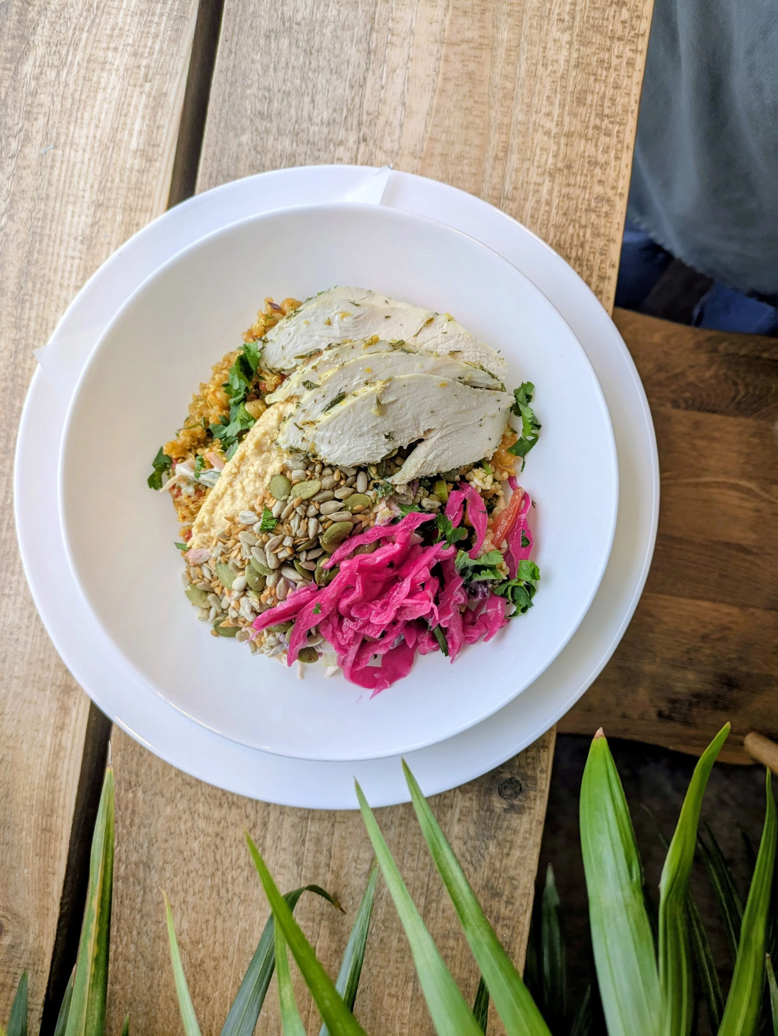 A white plate of sliced chicken breast, mixed grains, bright pink pickled cabbage, and chopped herbs, on a wooden table.