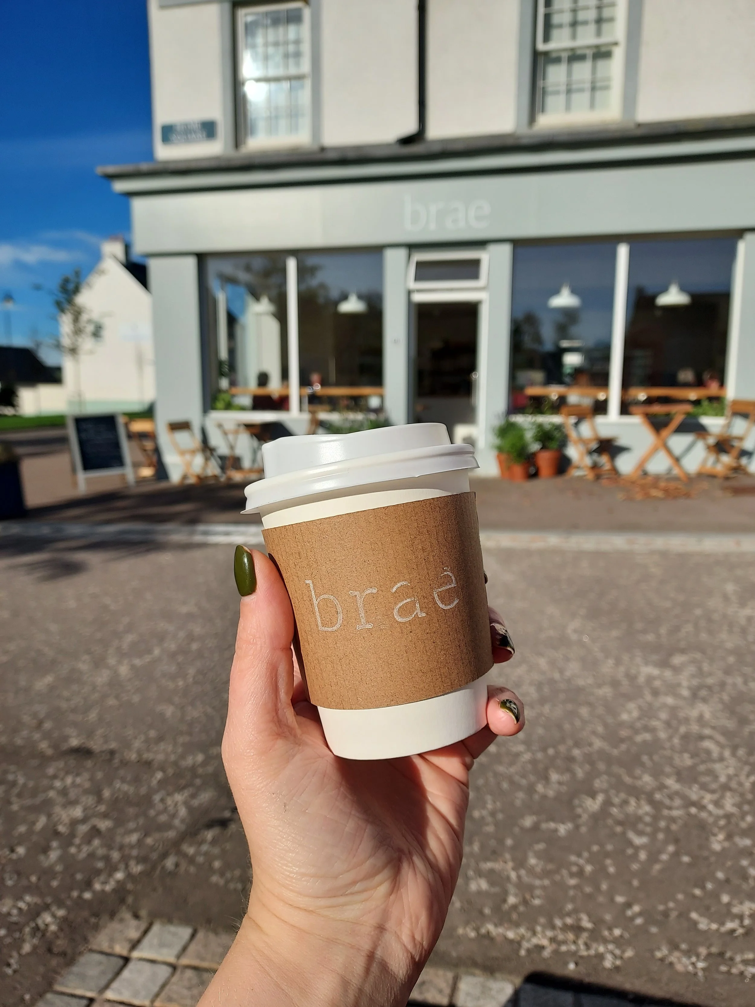 A person holding a to-go coffee cup outside a cafe named 'brae' on a sunny day.