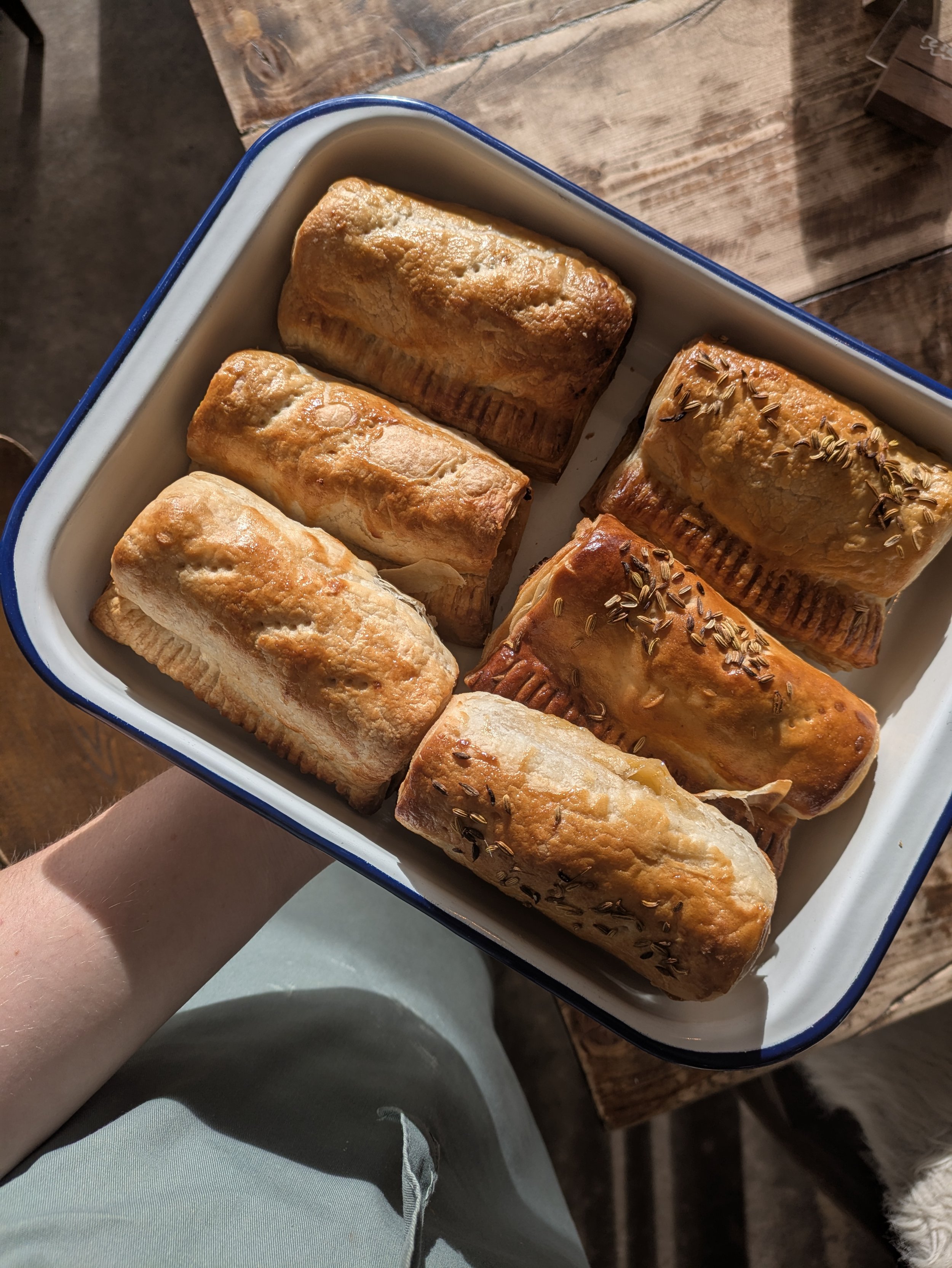 A hand holding a white dish filled with eight baked sausage rolls above a wooden table.  Local free range pork sausage made in house and baked each day.