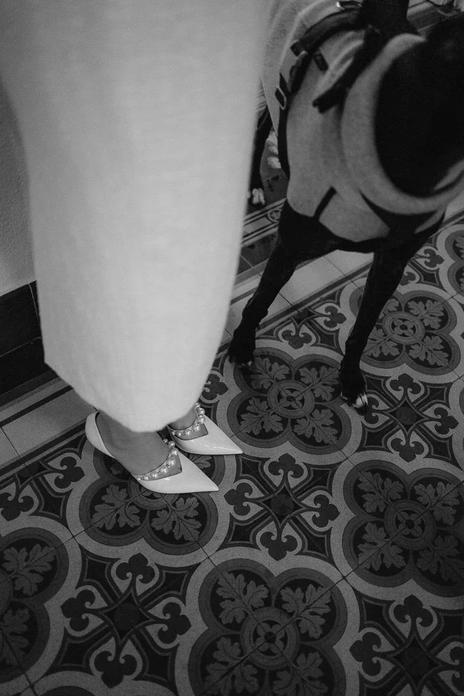 Authentische Hochzeitsfotografie in Bonn – Detailaufnahme der Brautschuhe mit Perlen und einem Hund auf gemustertem Boden in Schwarz-Weiß.