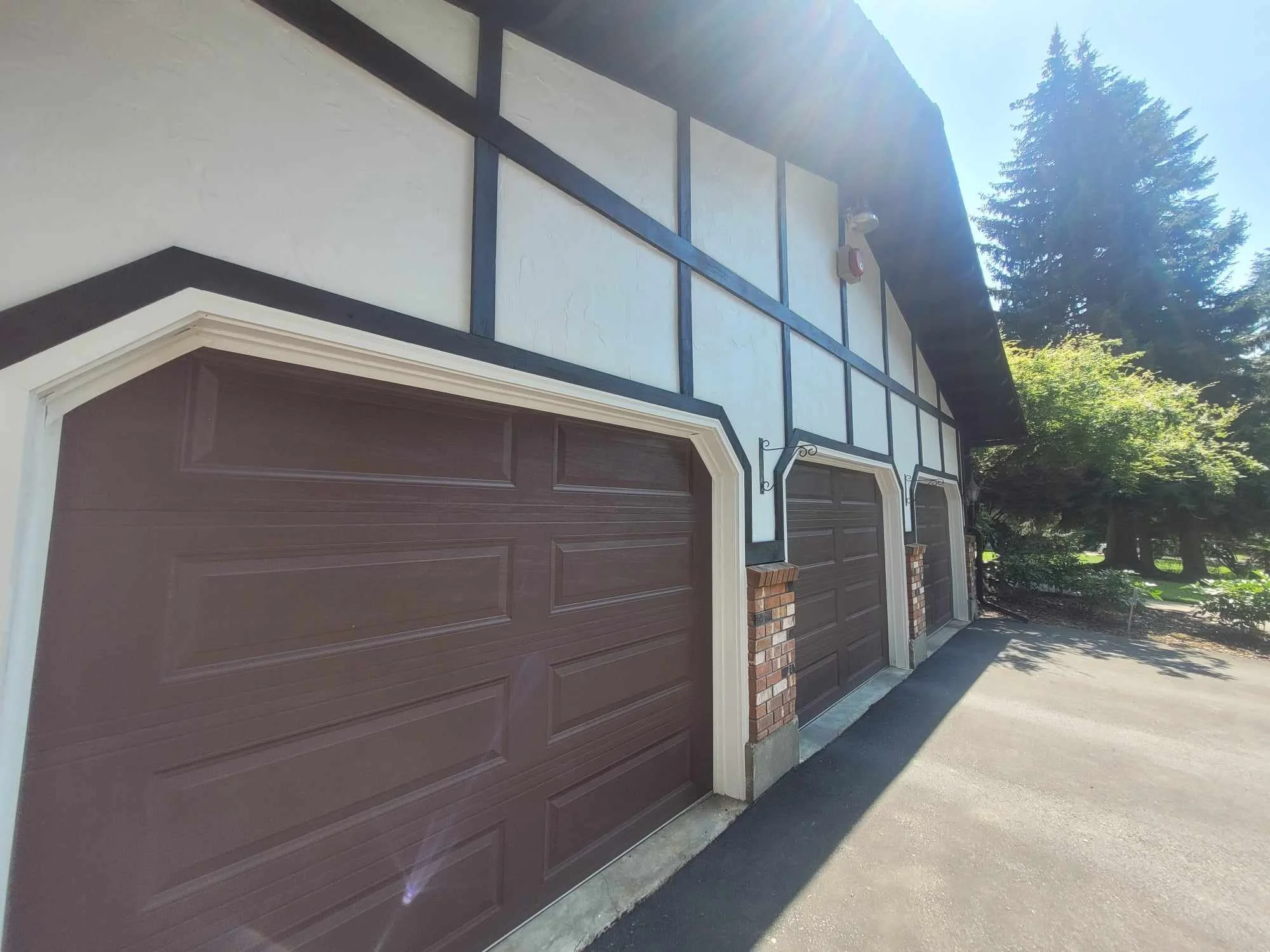 Image of a residential garage with three dark brown garage doors situated on a driveway. The house has a white exterior with dark trim details and brick accents at the bottom of the columns. Trees and greenery are visible in the background under bright sunlight.