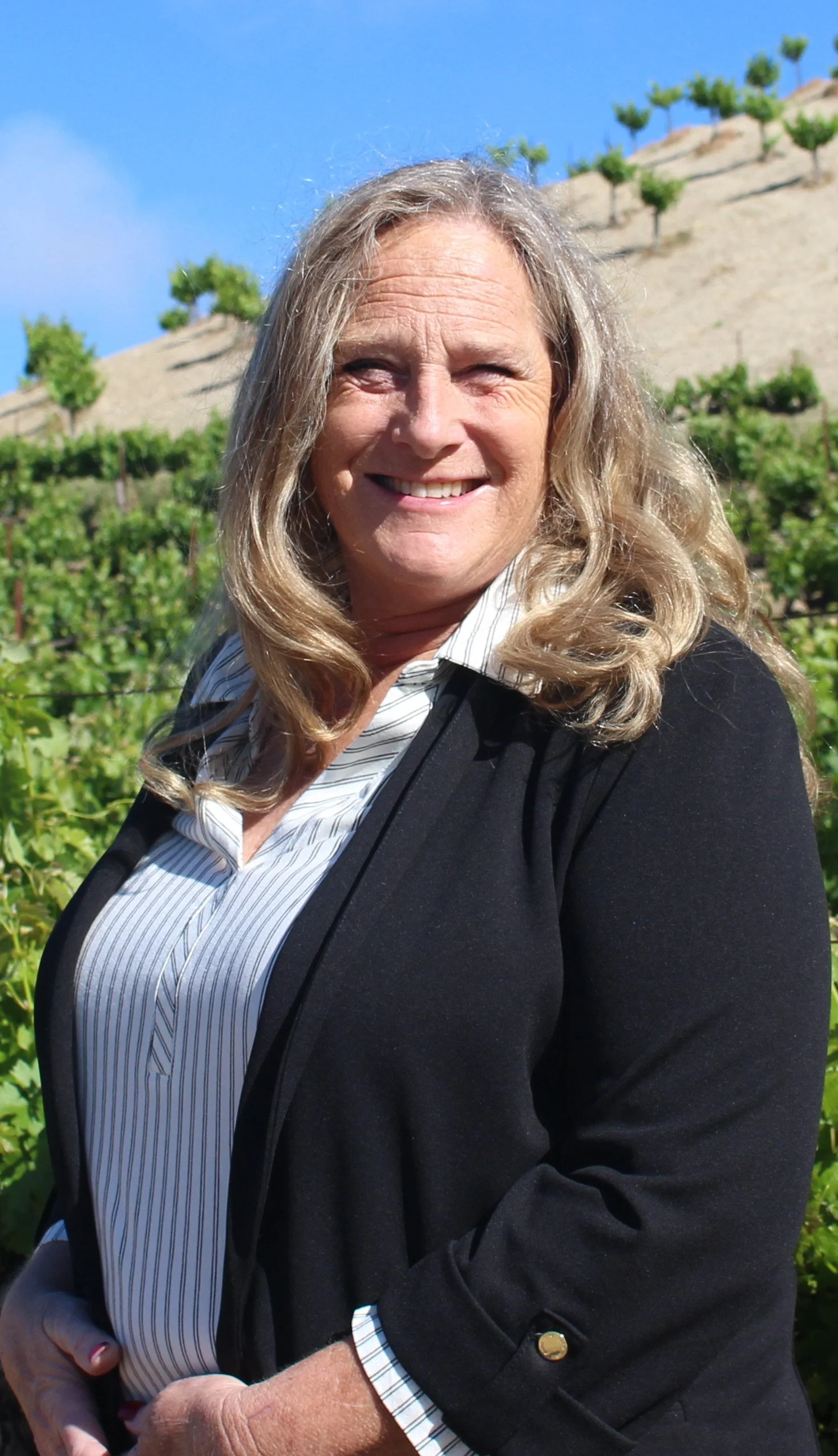 Tania with short blonde hair, wearing a black dress with eyelet pattern, standing in front of shelves of wine barrels in a winery or wine cellar.