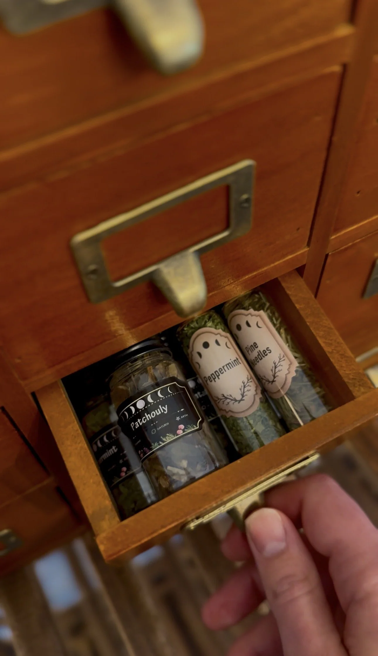 A person opening a small wooden drawer in a spice cabinet, revealing containers labeled 'Patchouli,' 'Peppermint,' and 'Cypress Needles' inside.