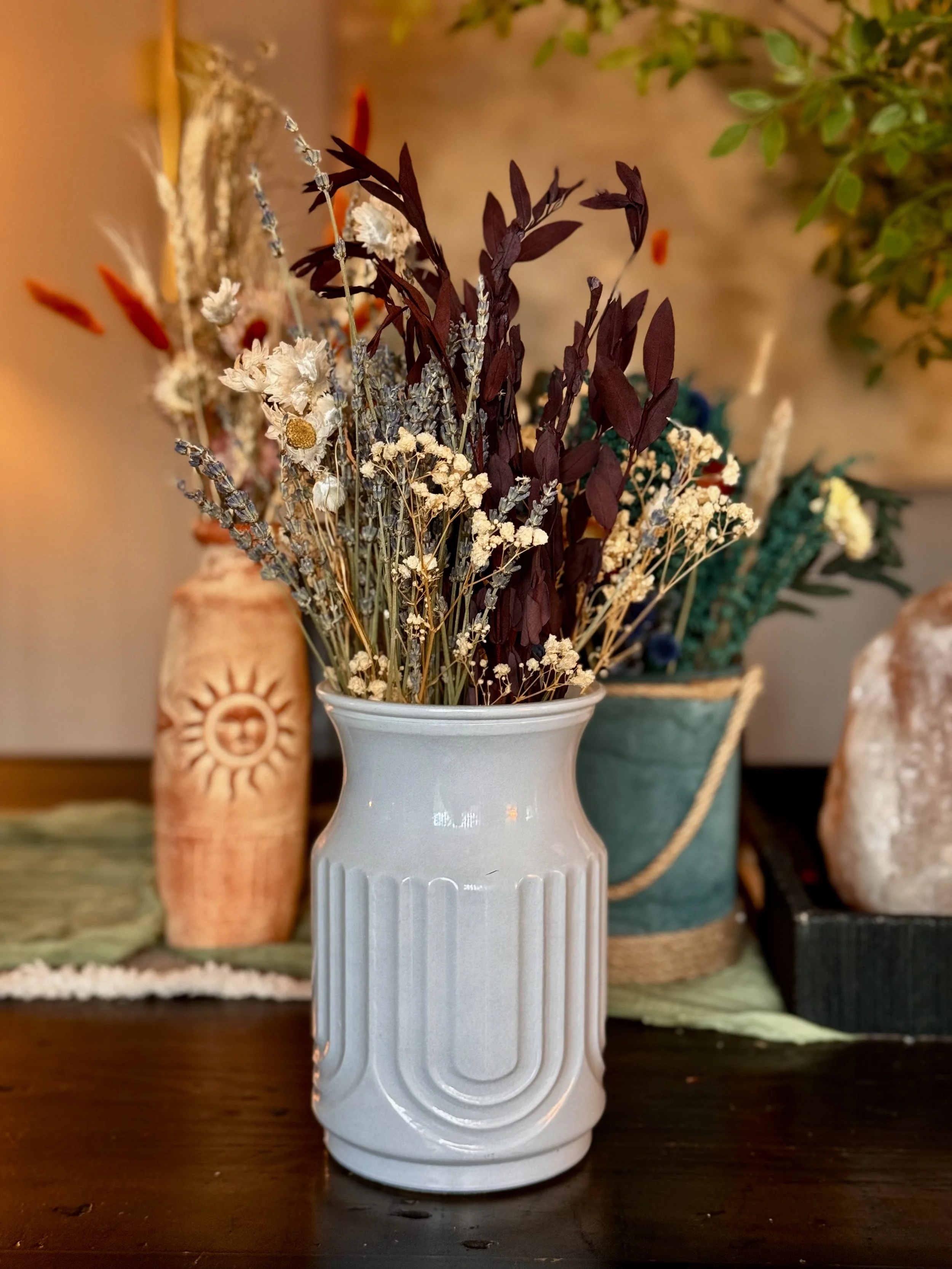A white ceramic vase filled with dried flowers and grasses, set on a wooden surface, with a blurred background of other containers and decor.