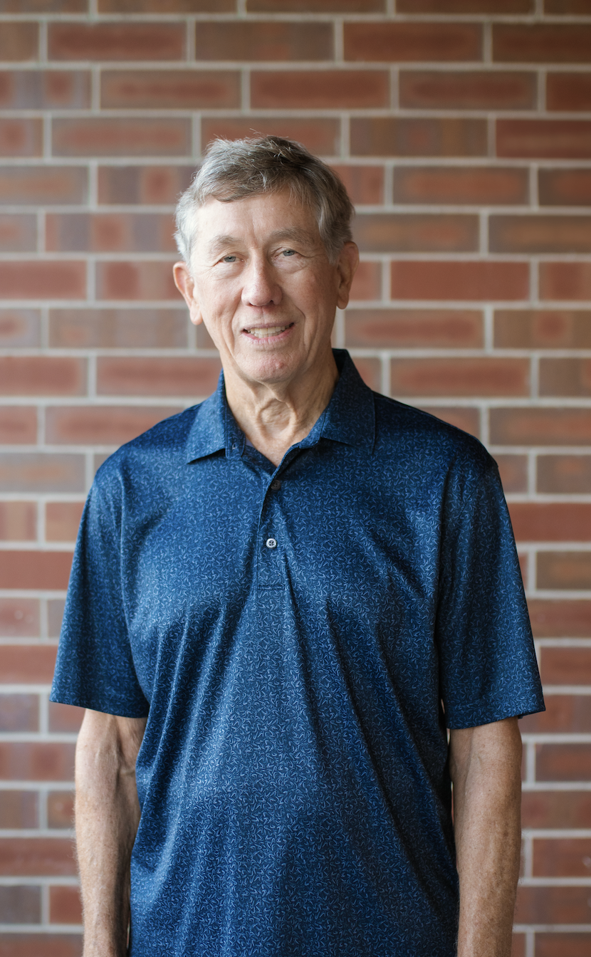 An older man wearing a blue patterned short-sleeved shirt stands in front of a brick wall.