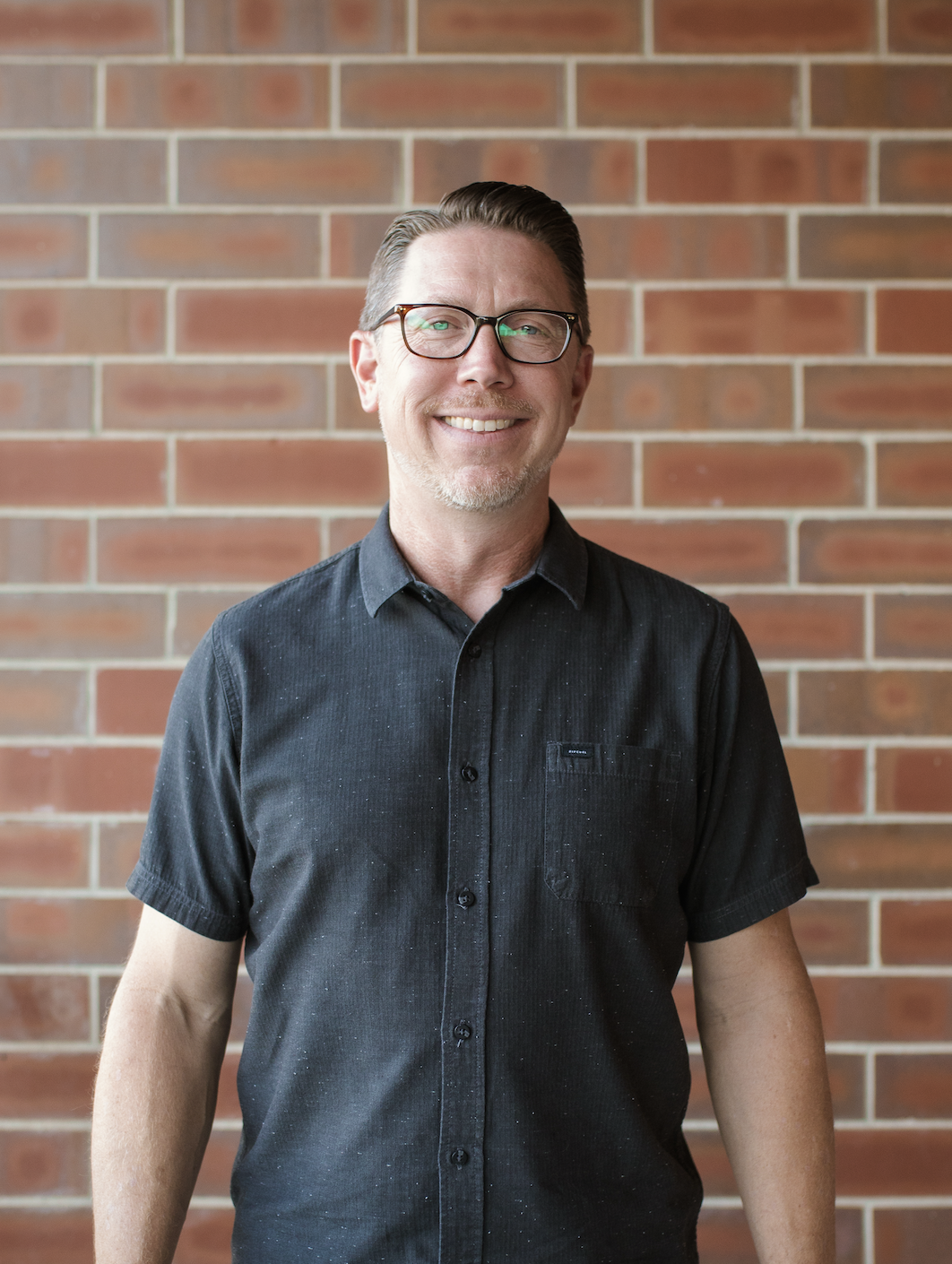 A man in glasses and a checkered shirt standing in a well-lit room with exposed brick and large windows.