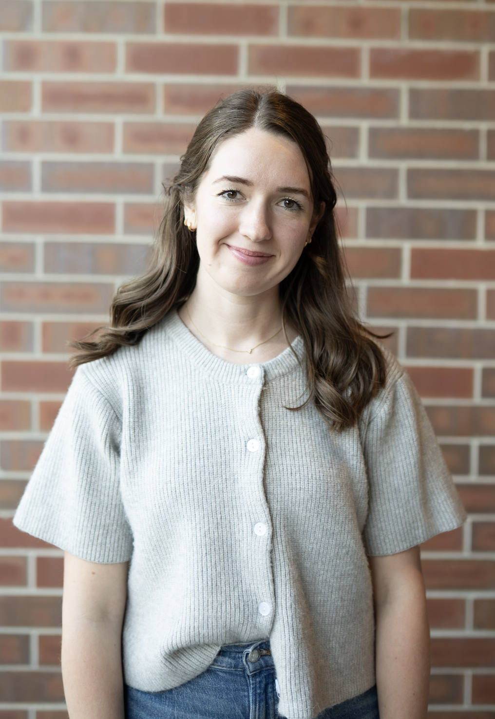 Young woman with brown curly hair smiling, wearing a denim dress, standing in front of a brick wall.
