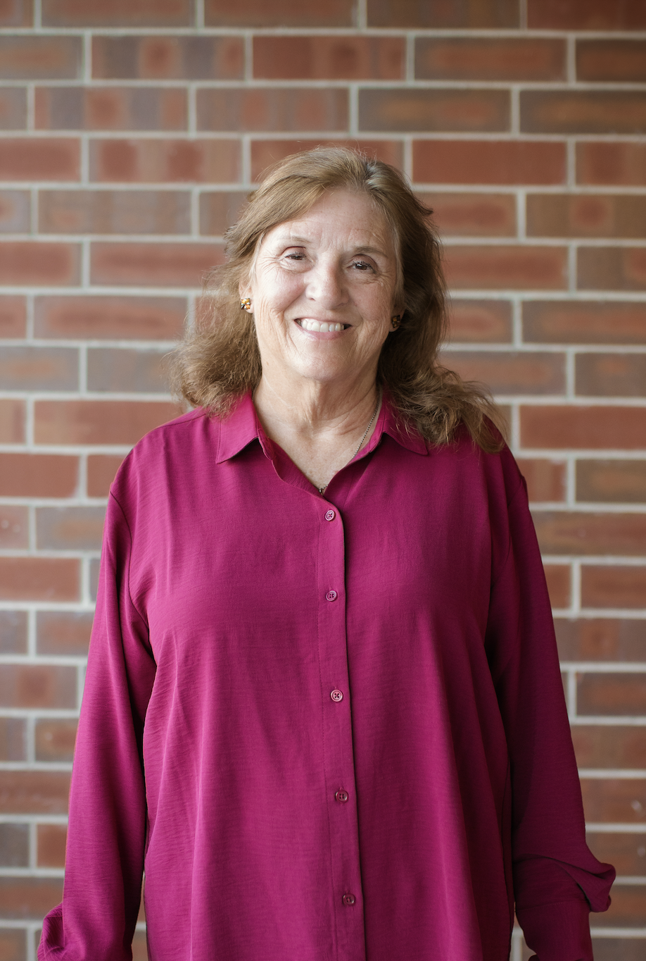 Smiling woman in a black polka dot shirt standing by a window inside a building with brick walls.