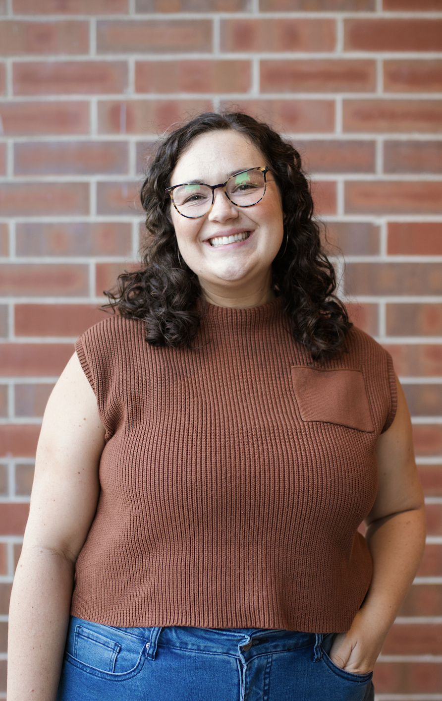A woman with curly dark hair and glasses smiling at the camera, standing in front of a brick wall.