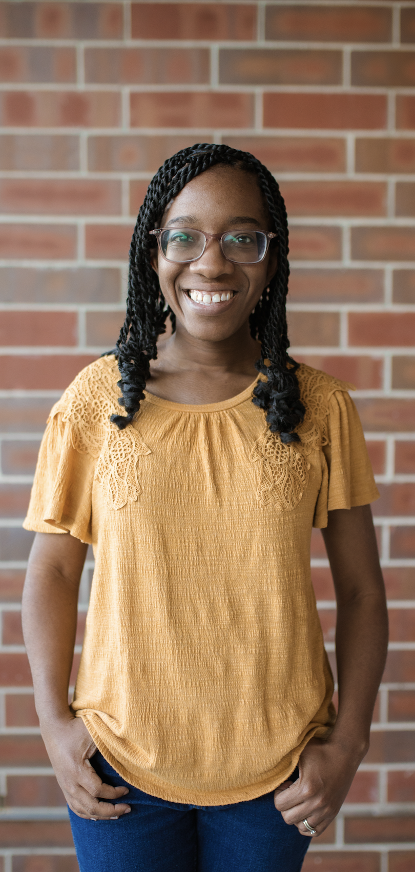 A young woman with glasses, braided hair, and a warm smile standing in front of a brick wall, wearing a mustard yellow blouse with lace details on the shoulders and blue jeans.