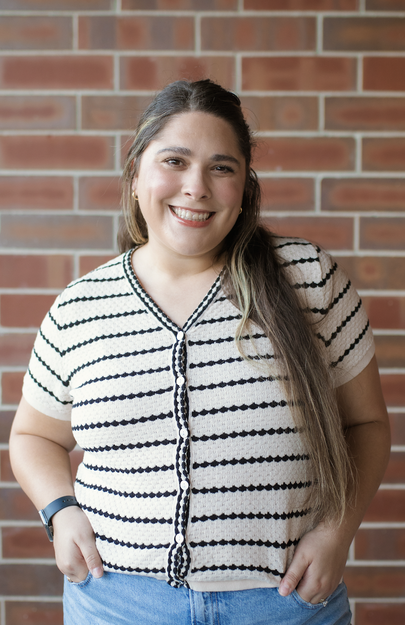 A young woman with long brown hair smiling, wearing a striped short-sleeve cardigan and blue jeans, standing in front of a brick wall.