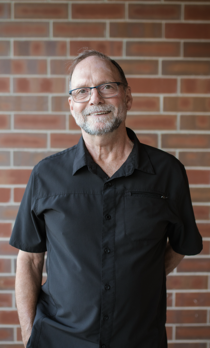 A smiling middle-aged man with glasses, a beard, and short hair, wearing a black button-up shirt, stands against a brick wall.