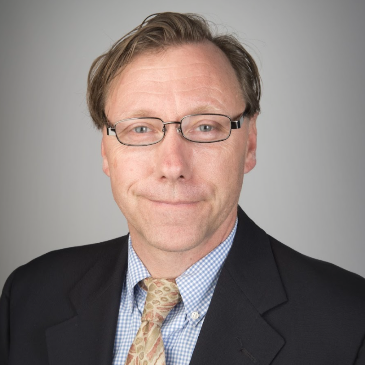 Headshot of a middle-aged man with light brown hair, glasses, wearing a checkered blue shirt, a patterned tie, and a black blazer against a plain gray background.