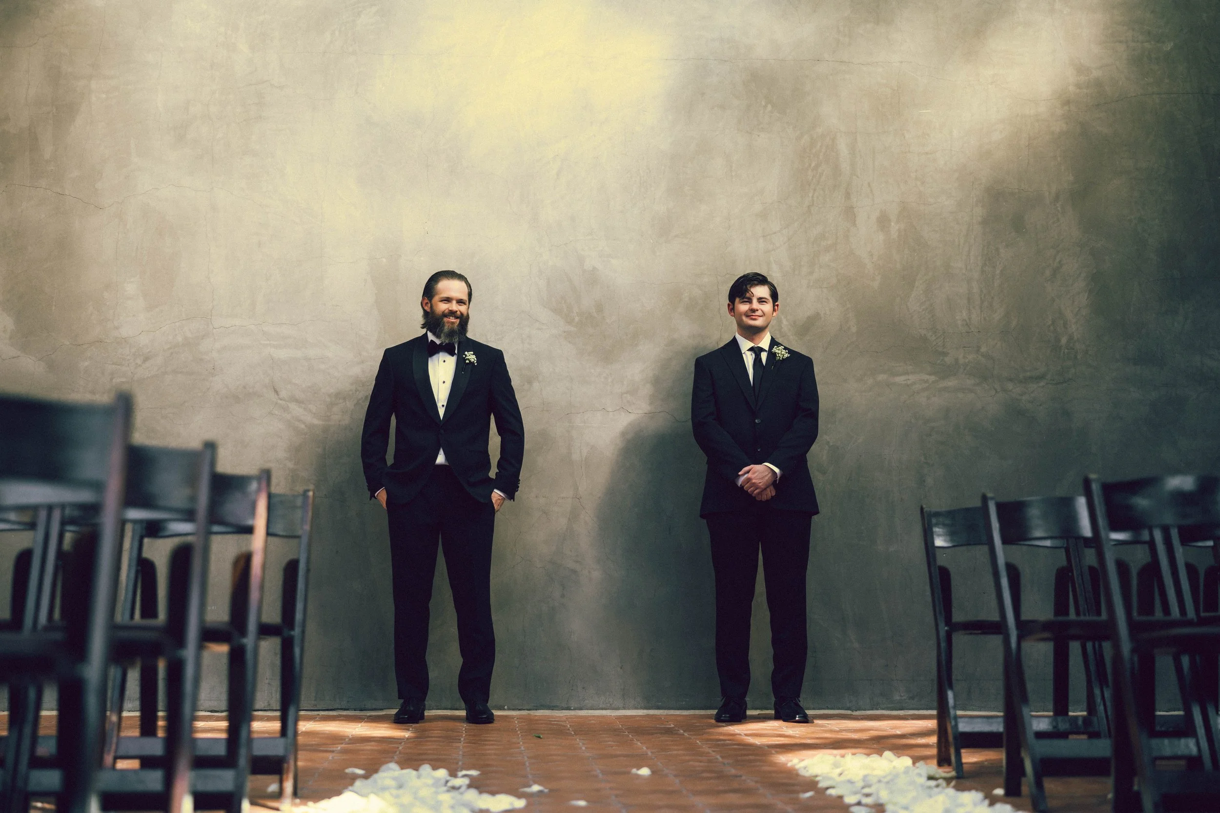 Two men dressed in tuxedos standing apart in an empty room with a large, textured gray wall behind them and chairs on either side, possibly for a wedding ceremony.