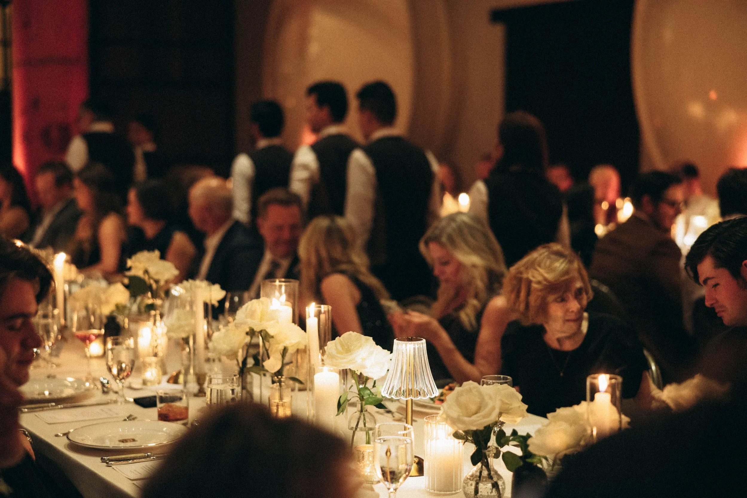 A dimly lit banquet hall filled with elegantly dressed guests seated at long tables decorated with white flowers, candles, and small lamps, in a warm, festive ambiance.