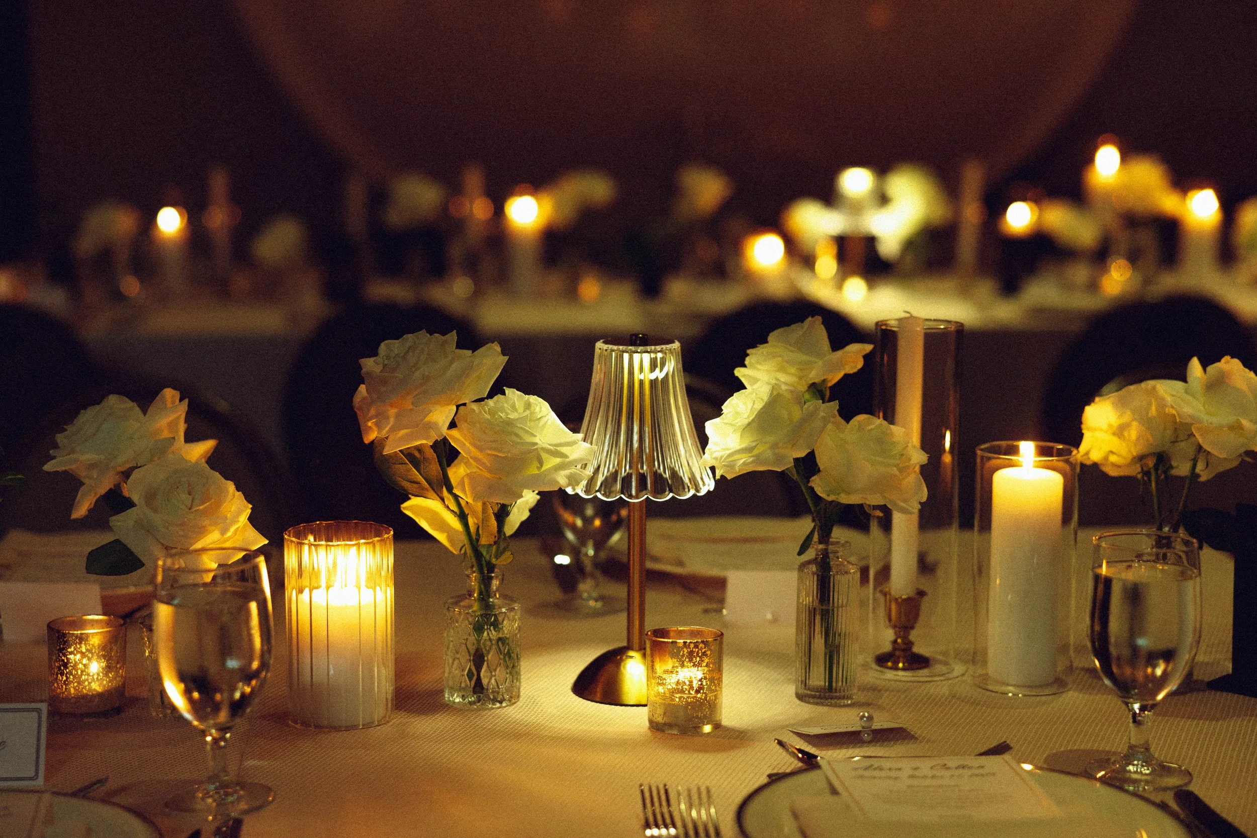 Elegant dinner table decorated with white roses in small vases, candles, and a small lamp with a pleated shade. Ambient lighting creates a warm and cozy atmosphere.