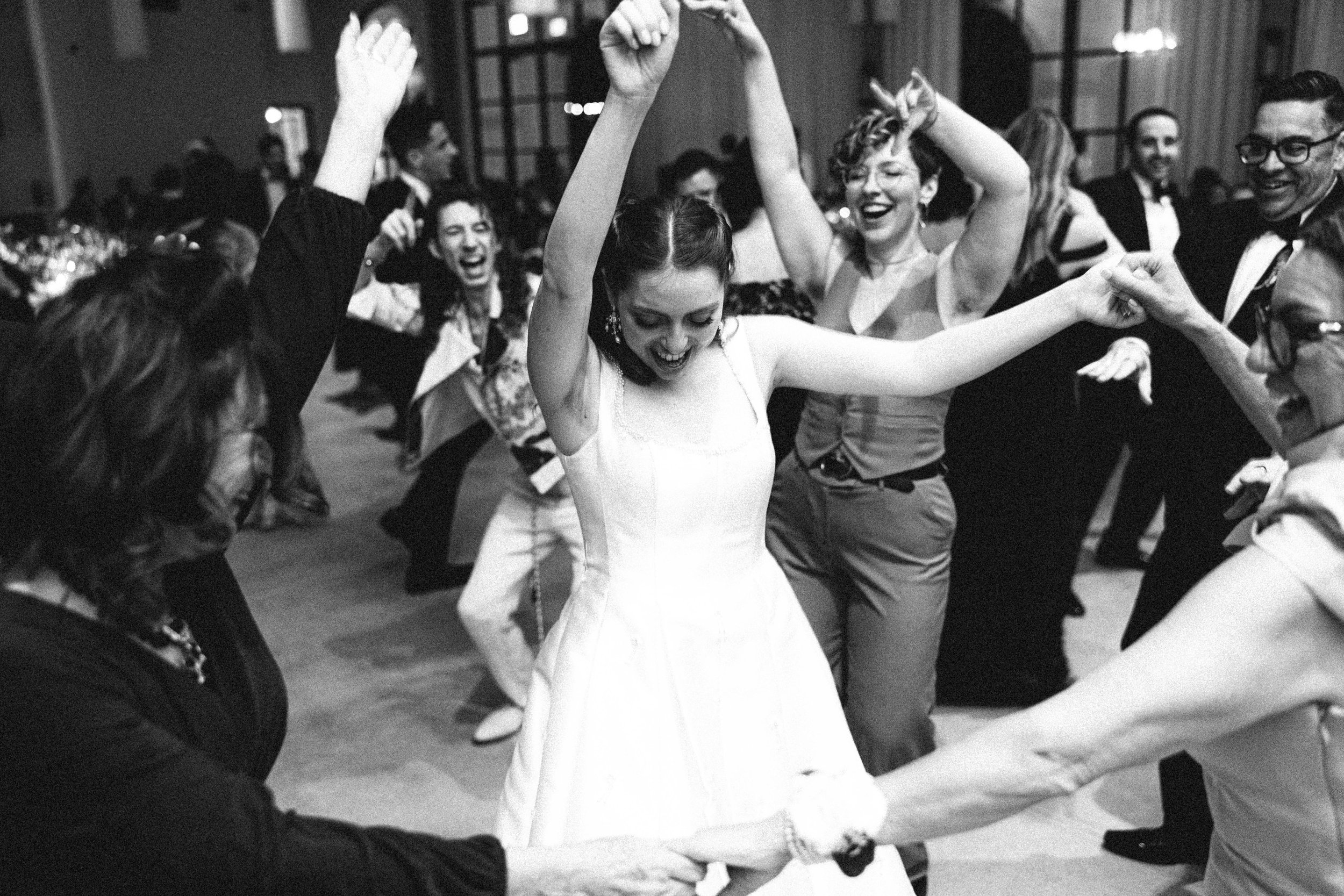 A group of people dancing and celebrating at a wedding reception, with a bride in a white dress in the center, holding hands with others, smiling and enjoying the moment.