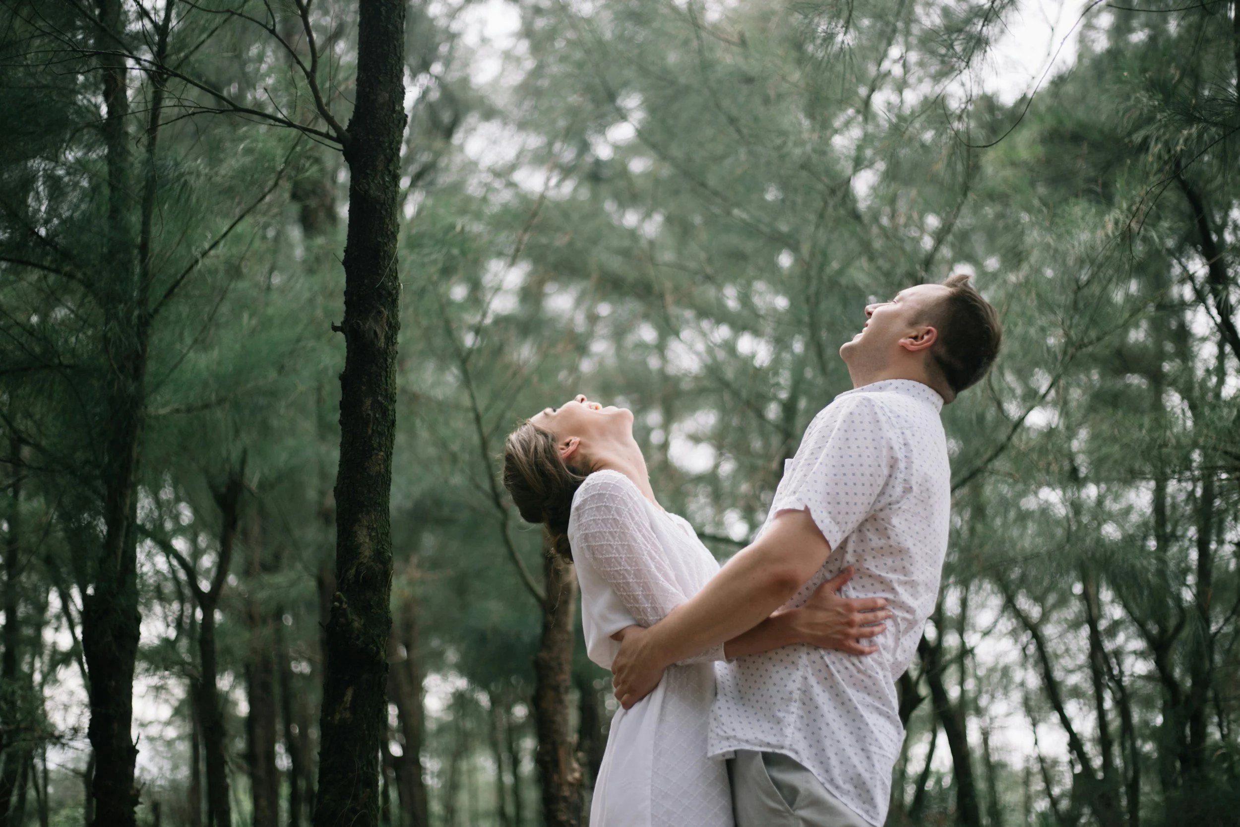 couple hugging, looking up to the tops of pine trees