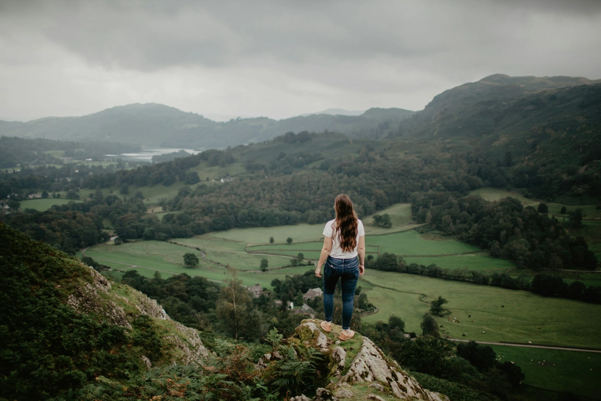 A woman stands on the moors overlooking the valley. She is wearing a white t-shirt and blue jeans.