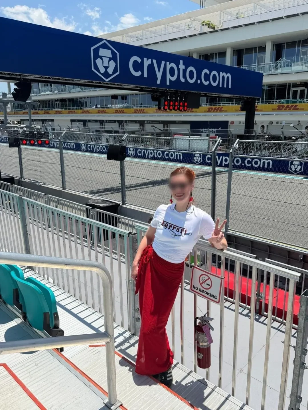 A woman with red hair wearing a white Ferrari t-shirt and red pants, posing and making a peace sign in a racing event area, with a sign indicating no smoking in front of her.