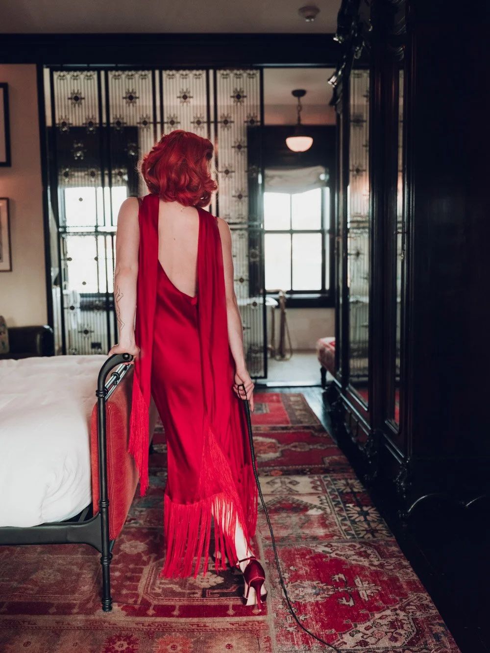New York Kinky Escort, Julia Flowerbomb, posing in a red silk dress, holding a leather whip