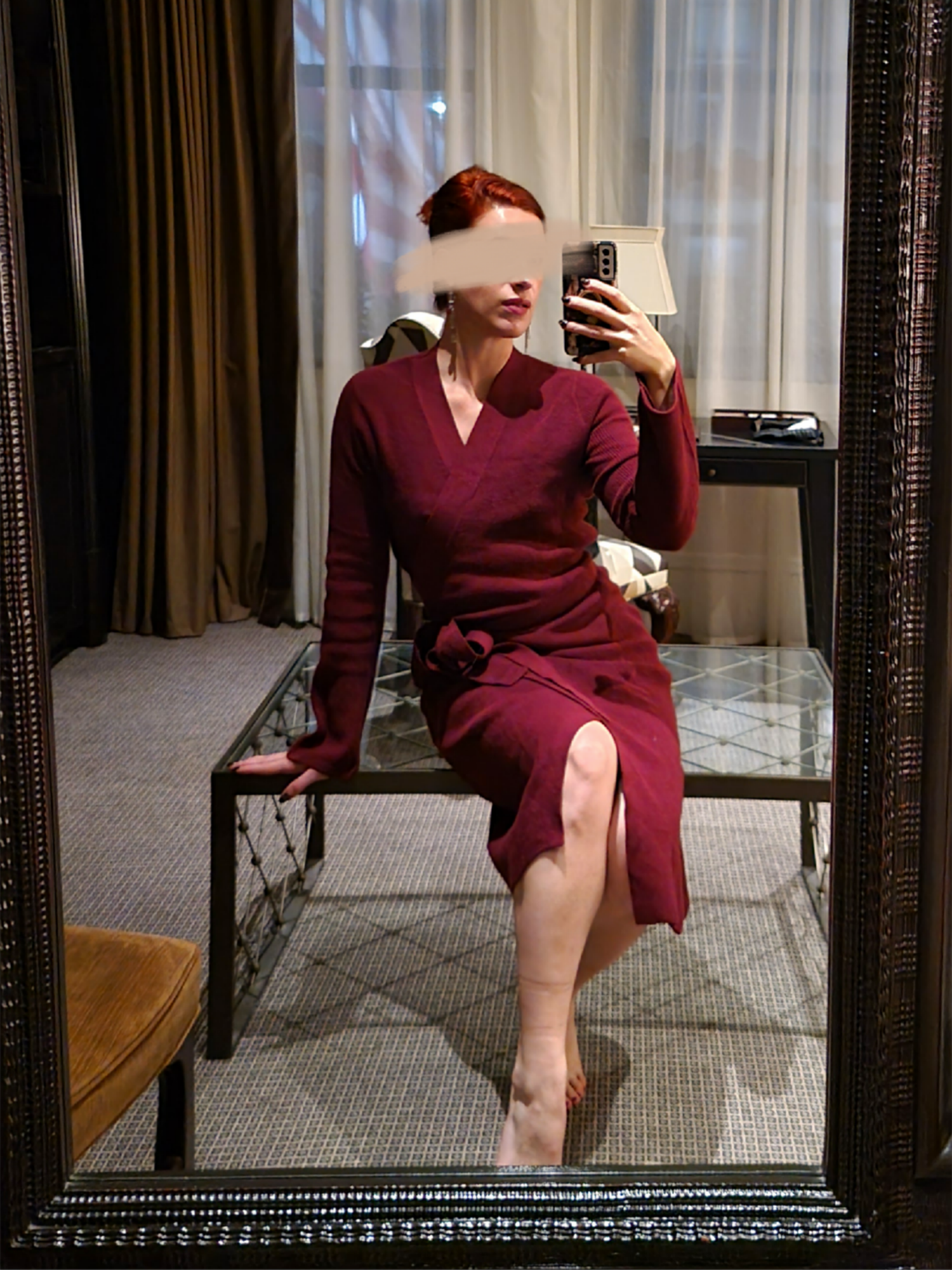 A woman with red hair taking a mirror selfie in a room with curtains and furniture, wearing a burgundy dress, with her face blurred for privacy.