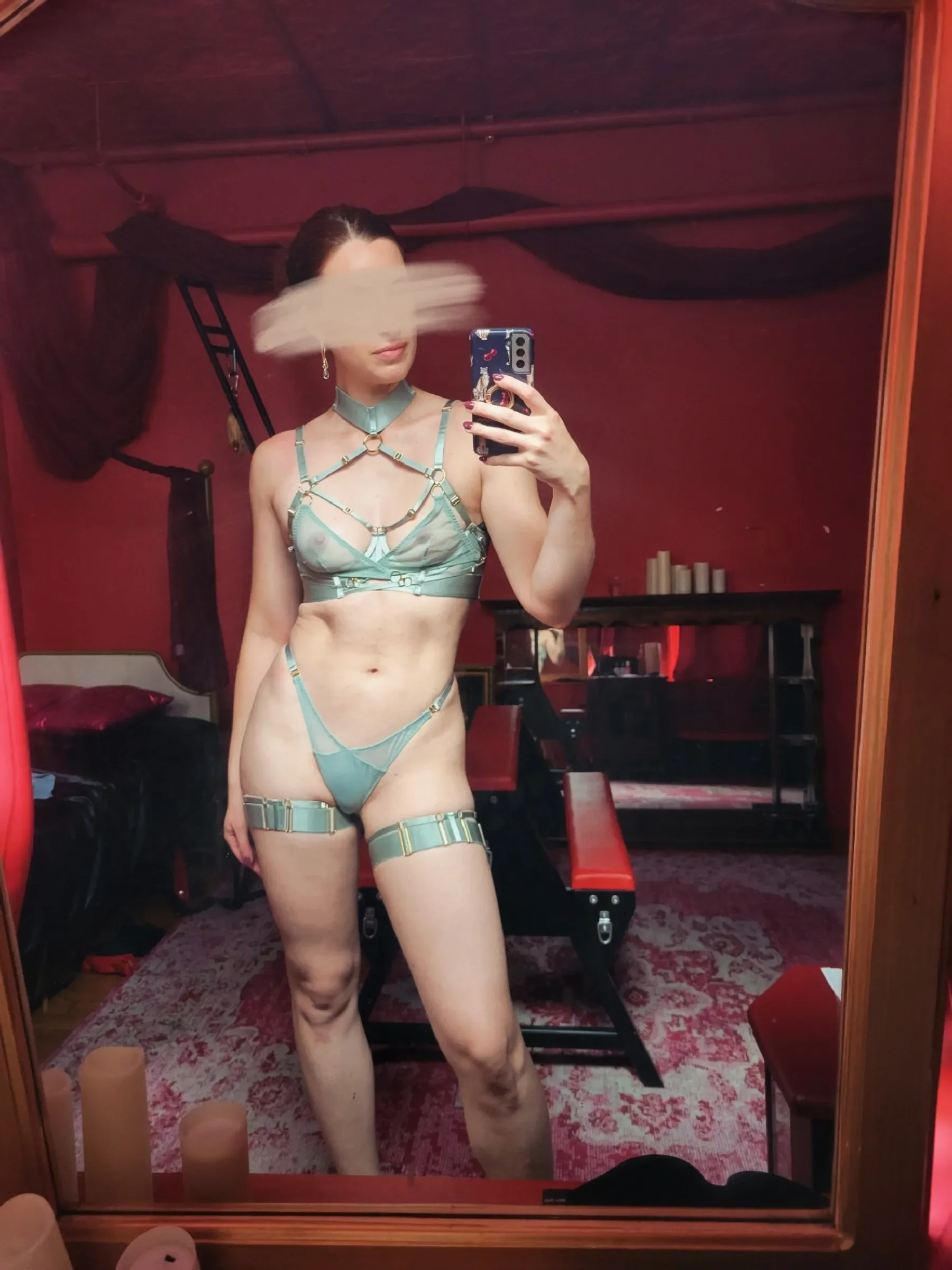 A woman taking a mirror selfie in a room, wearing latex and harness-style lingerie and a choker. Her face is blurred.