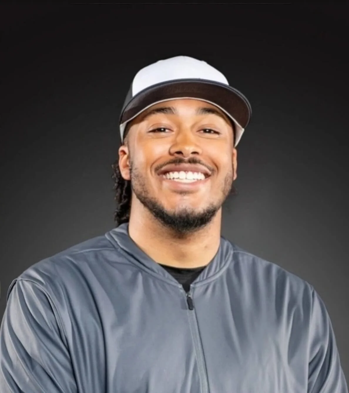 A smiling young man wearing a gray athletic jacket and a gray, black, and white baseball cap against a dark background.