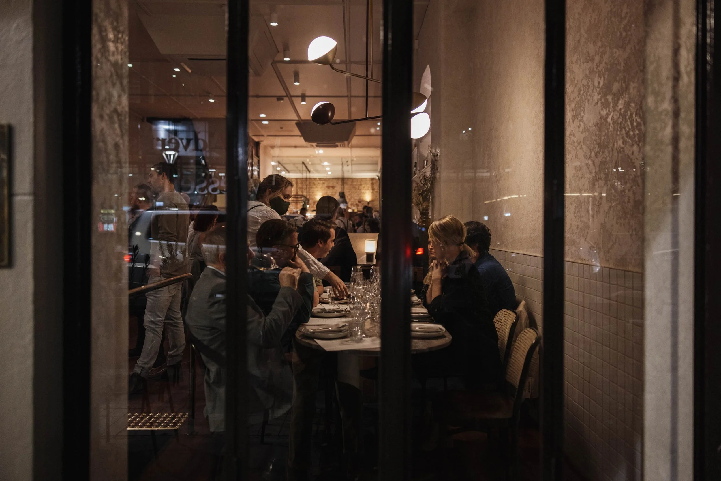 View of a restaurant interior through a window, showing people seated at a dining table, some talking and others serving, with warm lighting and modern decor.