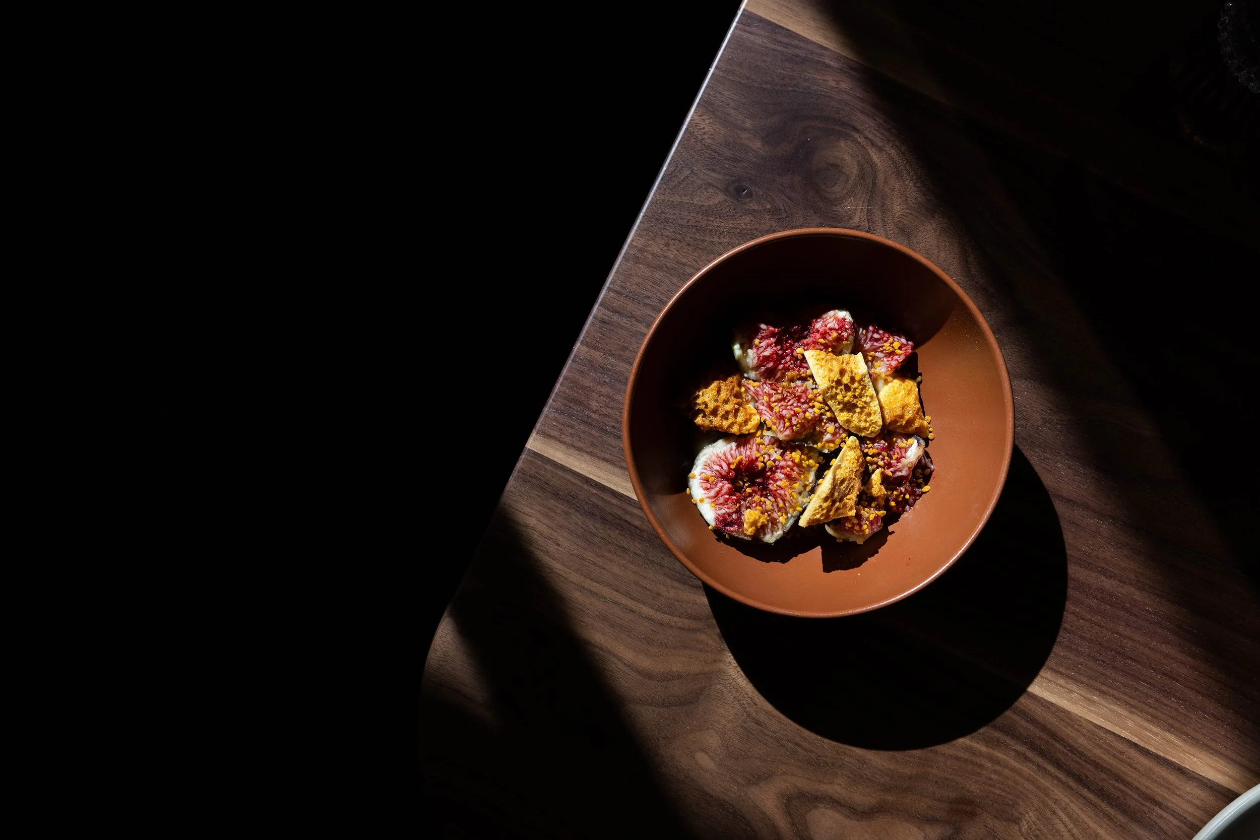 A brown bowl containing a dessert with figs and crispy toppings on a wooden surface, with light casting shadows.