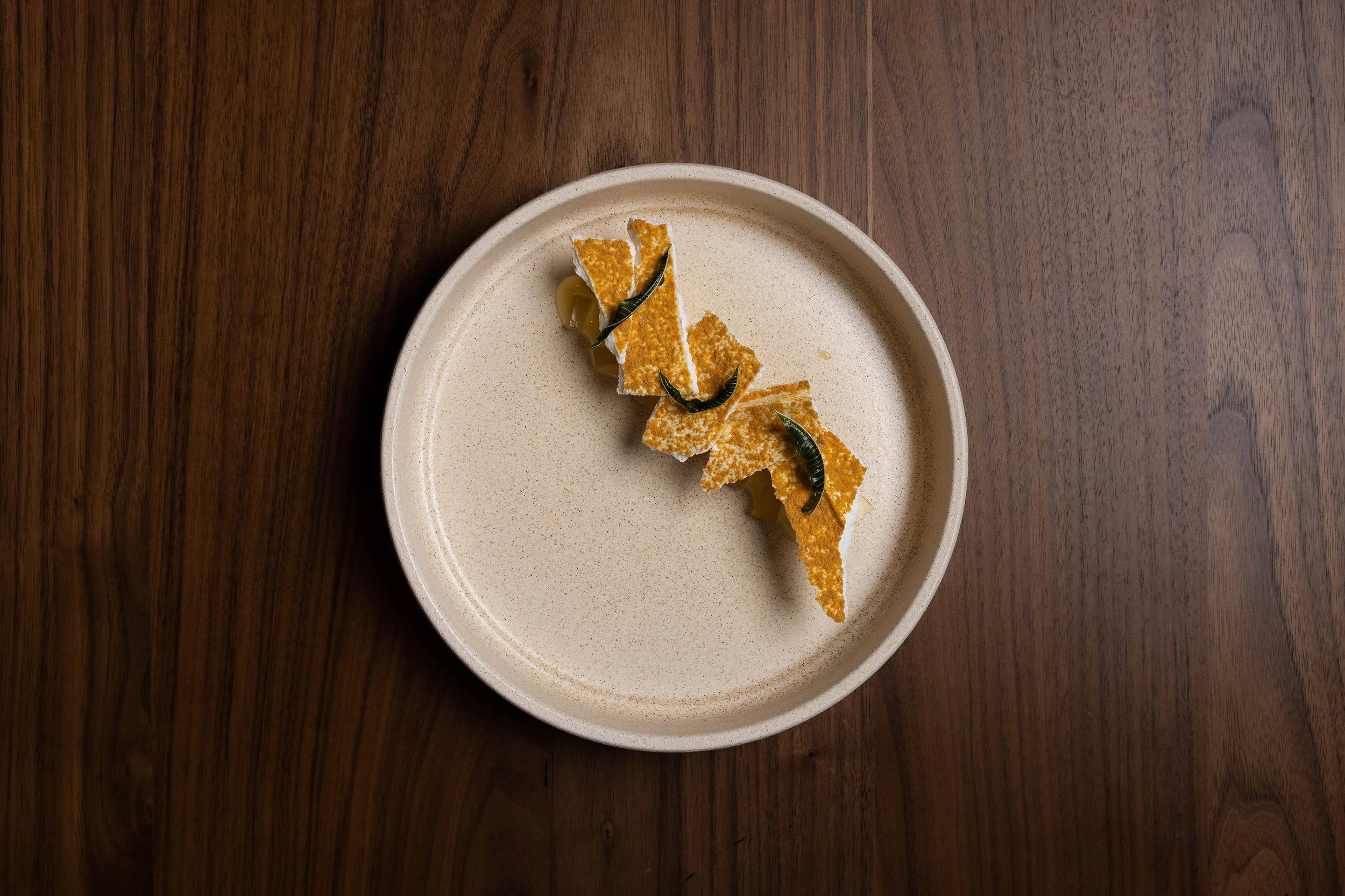 A round, beige plate with a small serving of sliced, crispy orange-colored food garnished with small green herbs, placed on a dark wooden surface.