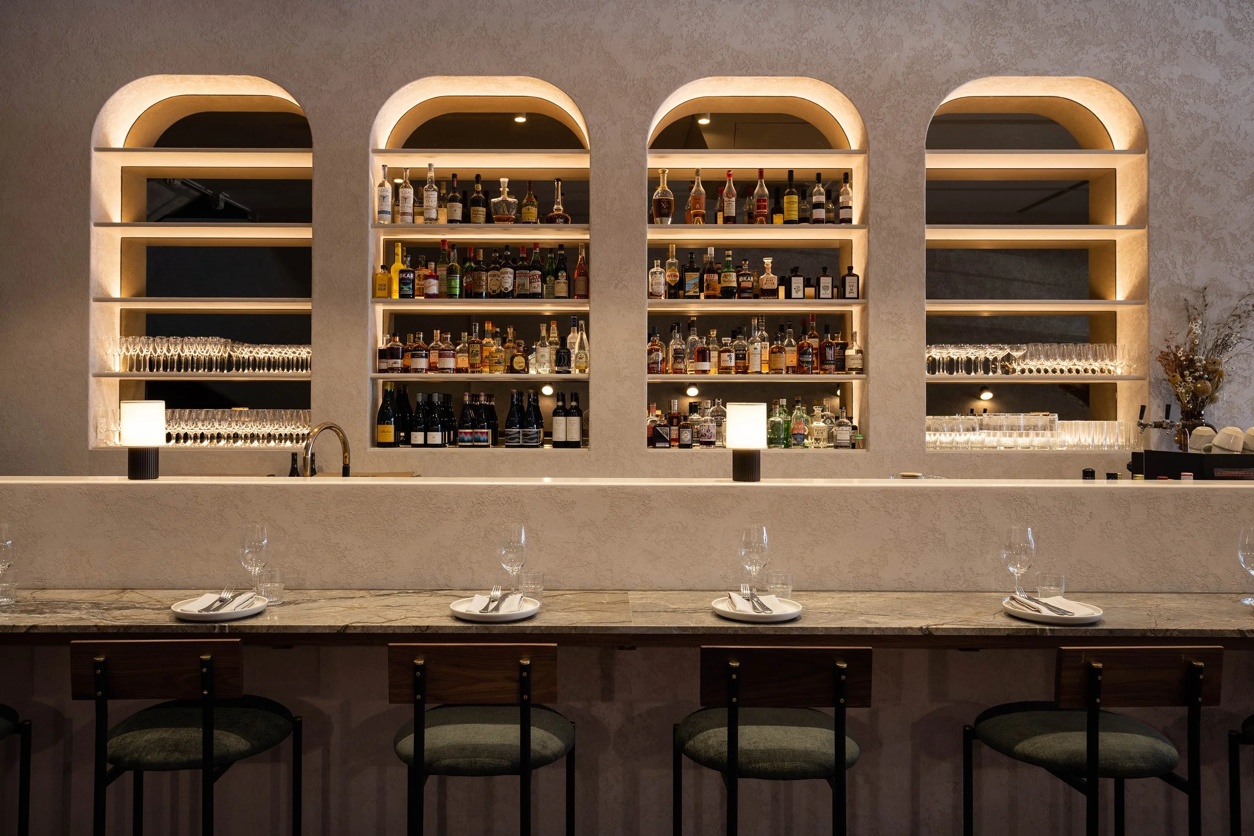 Bar counter with shelves filled with various bottles of liquor, illuminated with soft lighting, and set with glasses and plates in a modern, stylish restaurant.