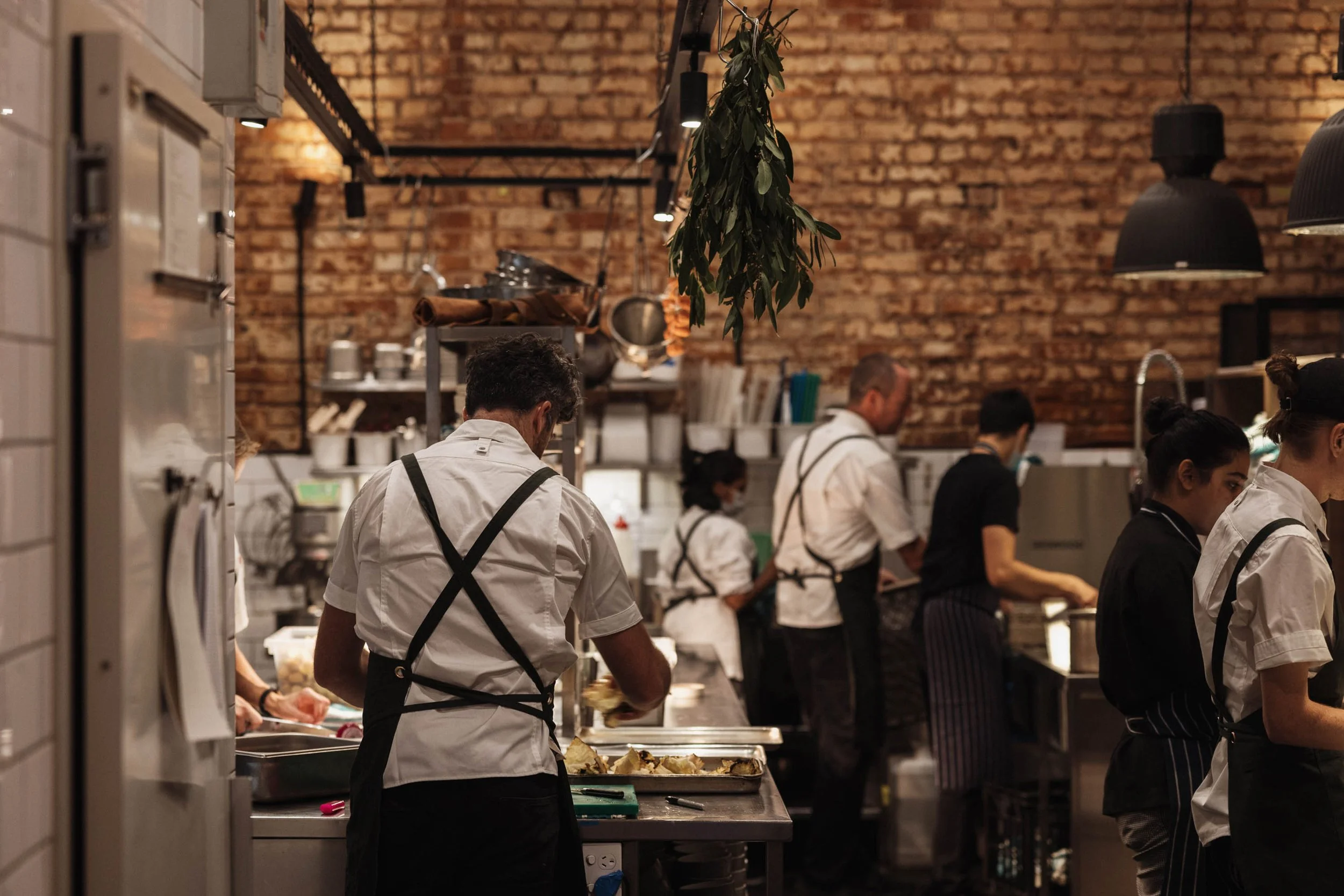 Kitchen staff working in a busy restaurant with an exposed brick wall and hanging herbs.