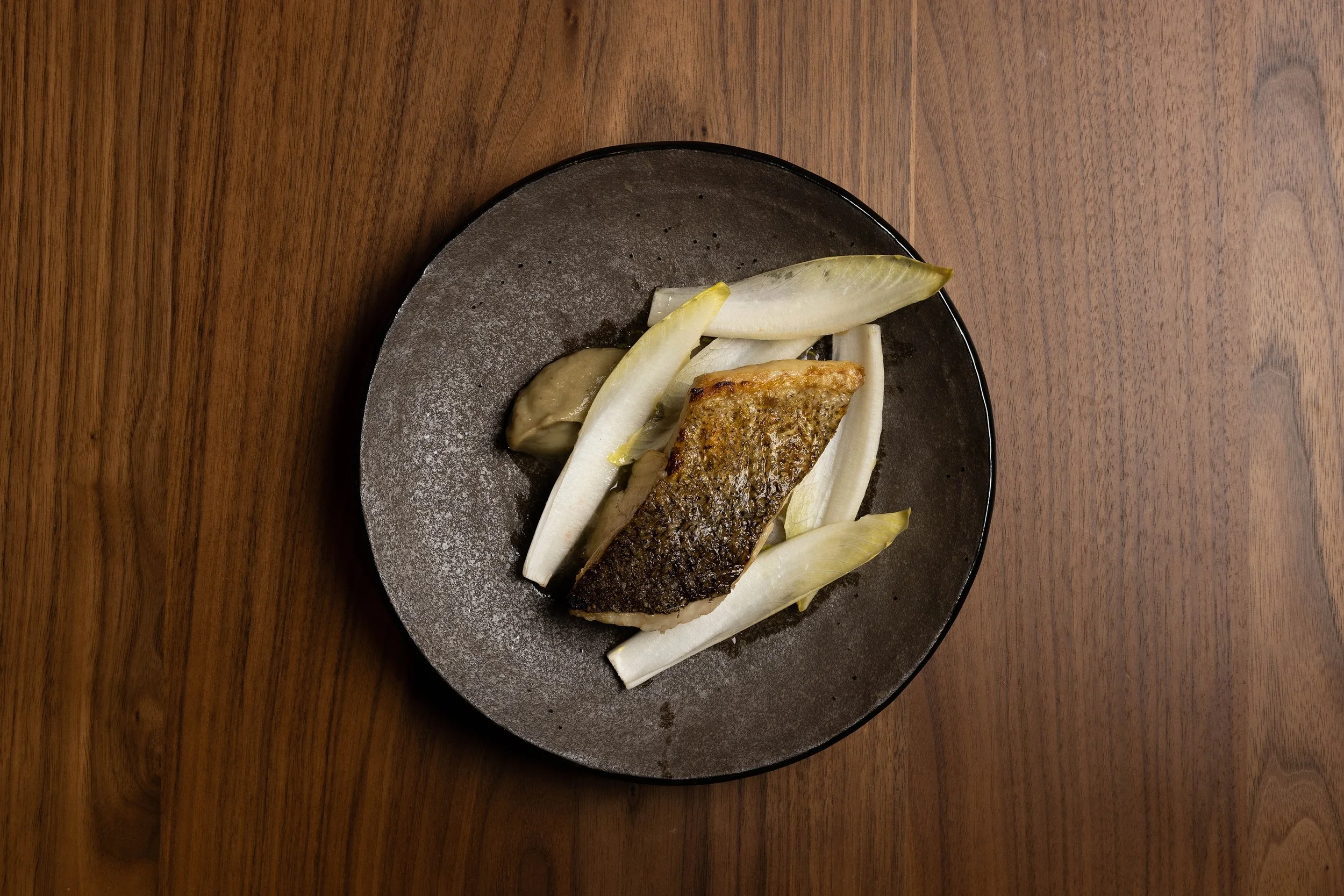 A black plate with a seared fish fillet, accompanied by white and pale green vegetables, on a wooden surface.