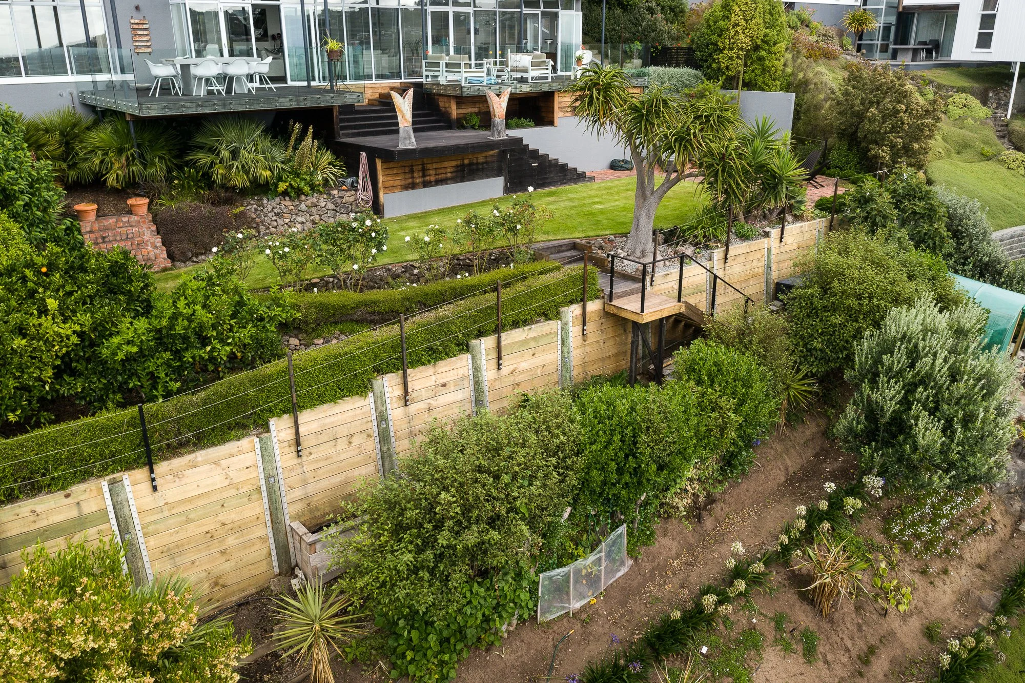 A modern backyard garden with a multi-level deck, lush green grass, trees, bushes, and a garden area with plants and shrubs, with an outdoor dining table and chairs on the upper deck.