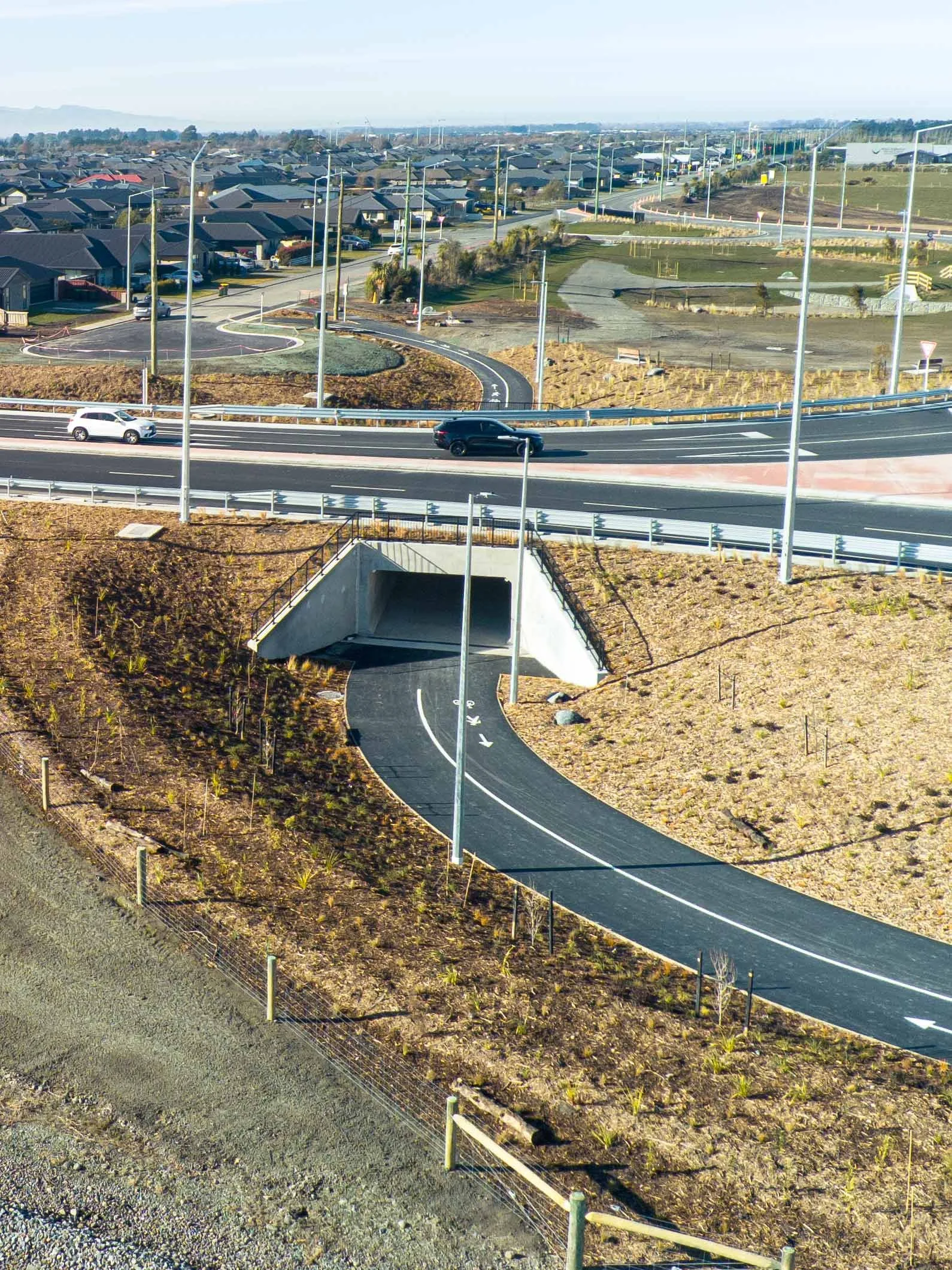 A view of a suburban neighborhood under construction with new roadwork and landscaping, featuring a curved road, streetlights, and residential houses in the background.
