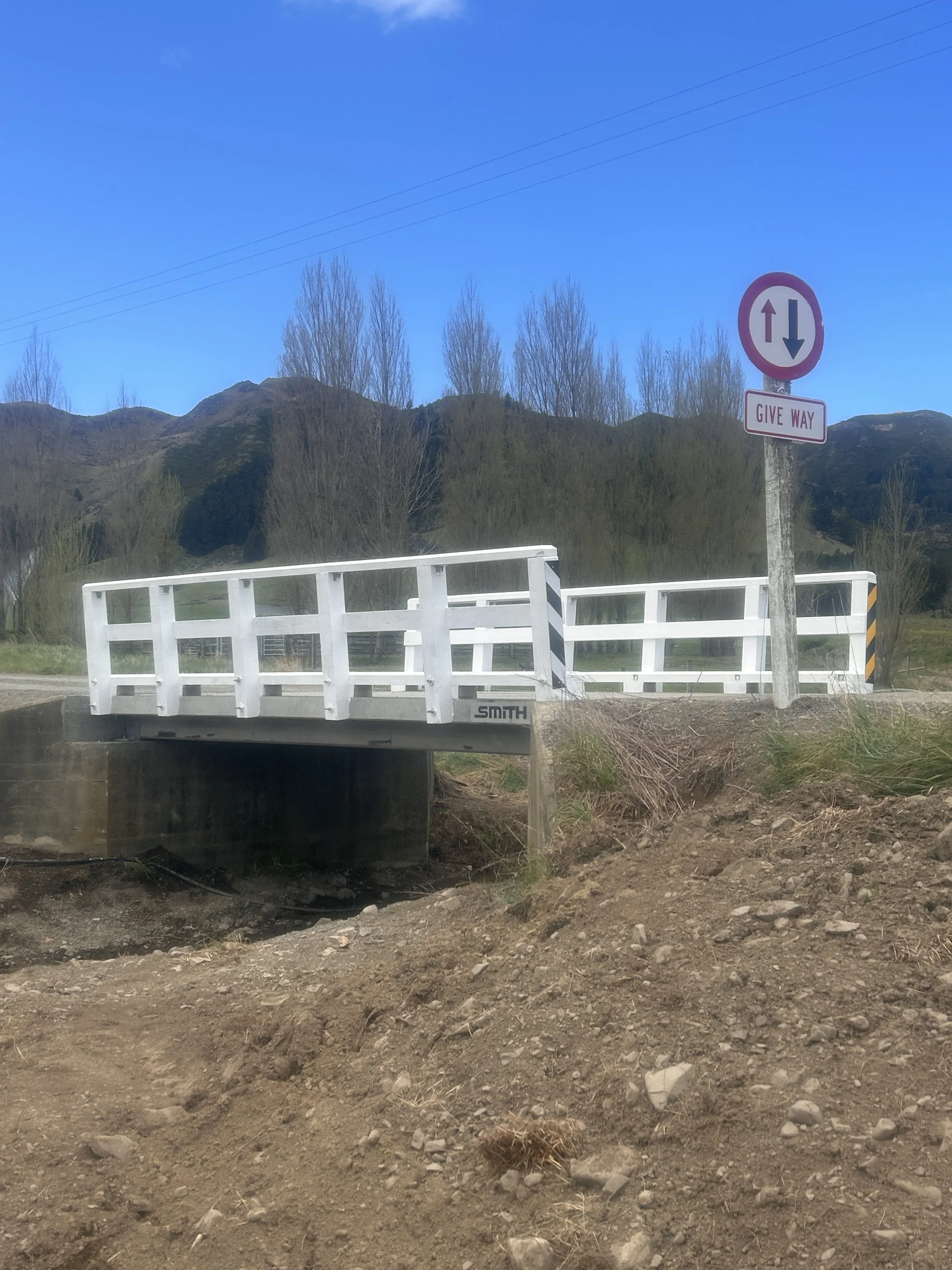 A white bridge with black and yellow striped markings on the sides crosses over a small drainage or culvert. To the right, a circular traffic sign with arrows indicating two-way traffic and a sign below that reads 'GIVE WAY'. In the background, there are leafless trees, mountains, and a clear blue sky.