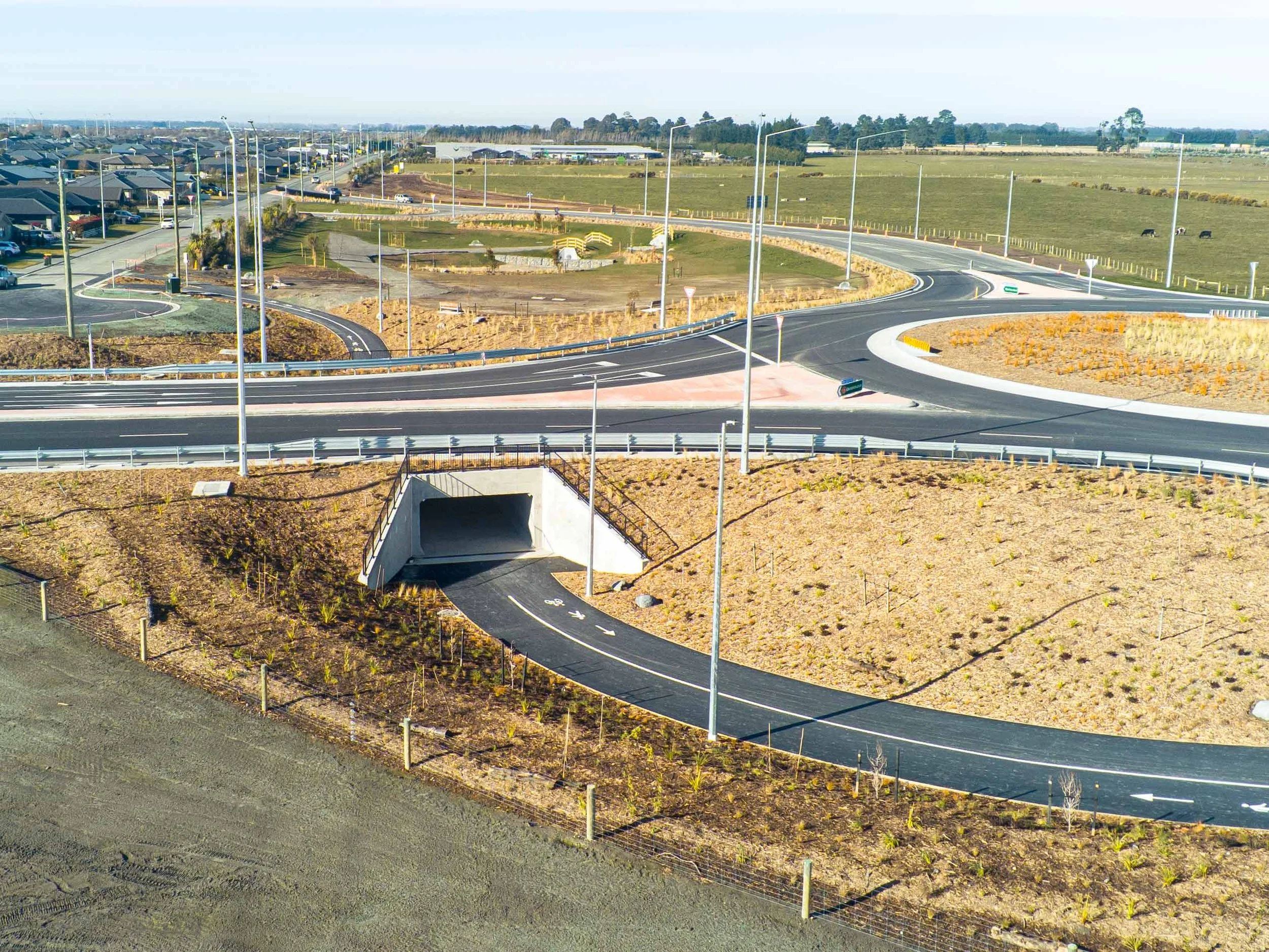 Construction site with curved paved roads, streetlights, and a view of nearby residential houses and open fields