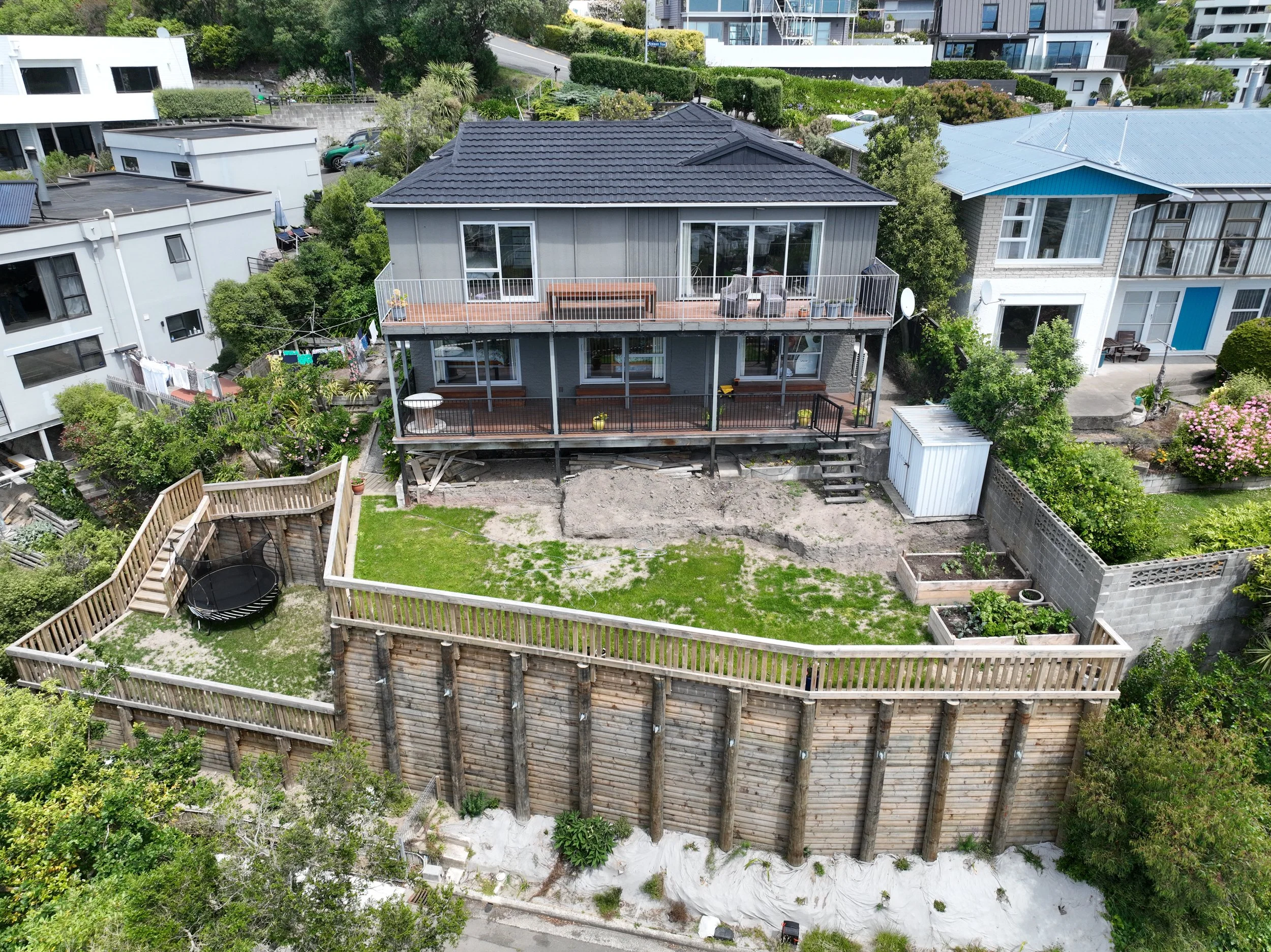 An aerial view of a multi-story gray house with a dark-colored roof and large windows, featuring two spacious balconies with outdoor furniture. The house is surrounded by lush greenery, neighboring houses, and a backyard with a grass lawn, a fenced-in garden area, and a trampoline.