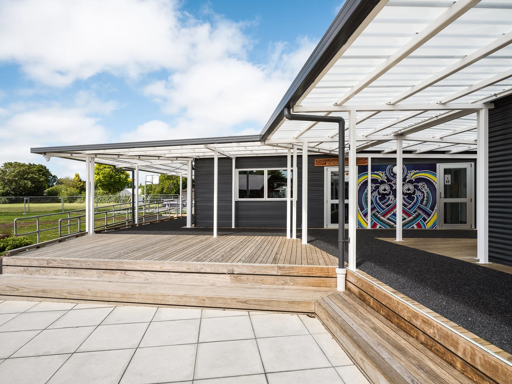 An outdoor area with a wooden deck and a tiled section, featuring a modern building with a black and gray exterior, white framing, a colorful mural on the wall, and a staircase leading up to a covered area with a metal roof. Green trees and a blue sky with clouds are visible in the background.