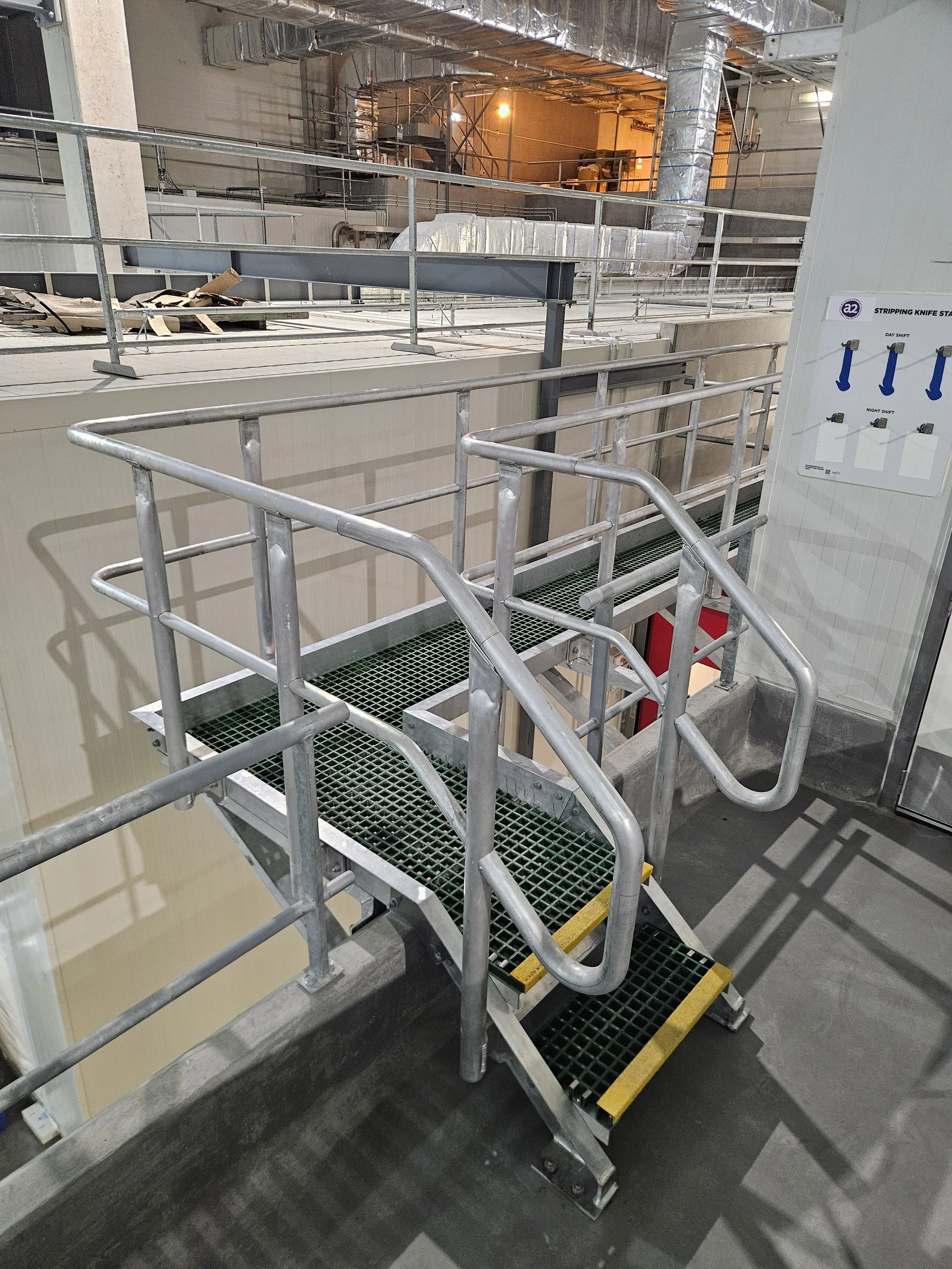 Metal staircase with handrails, leading to an industrial workspace with piping and machinery in the background. The area is lit with warm and cool lighting.
