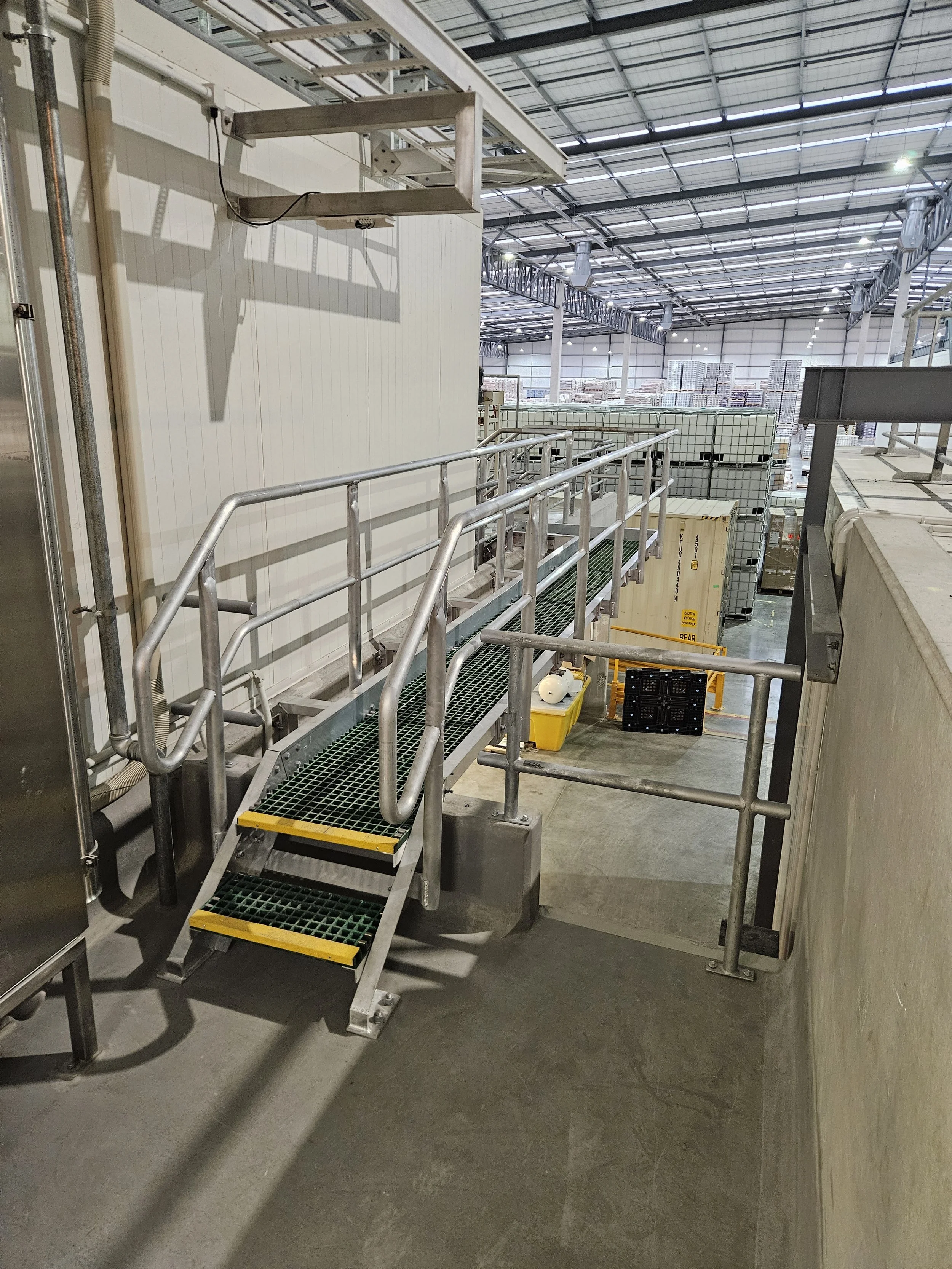 Industrial warehouse with metal stairs, railings, and conveyor systems for material handling, with stacked pallets in the background.