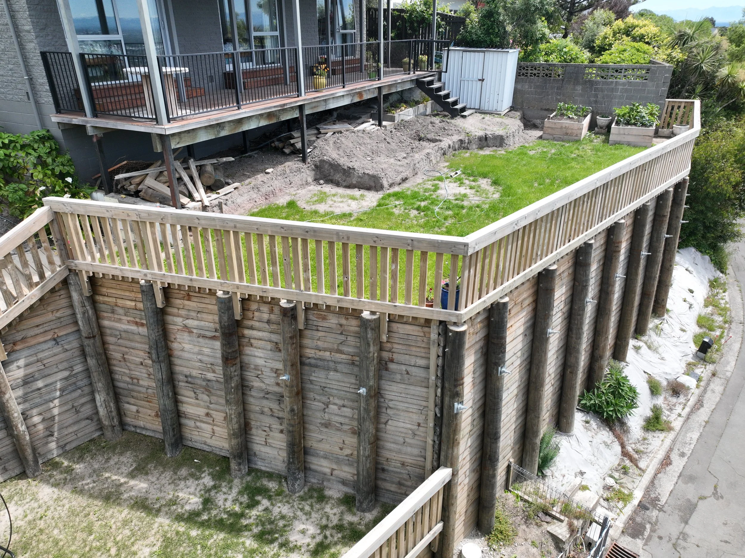 Backyard with a deck under construction, showing exposed dirt, wood debris, and building materials, with a green lawn, trees, plants, and neighboring buildings in the background.