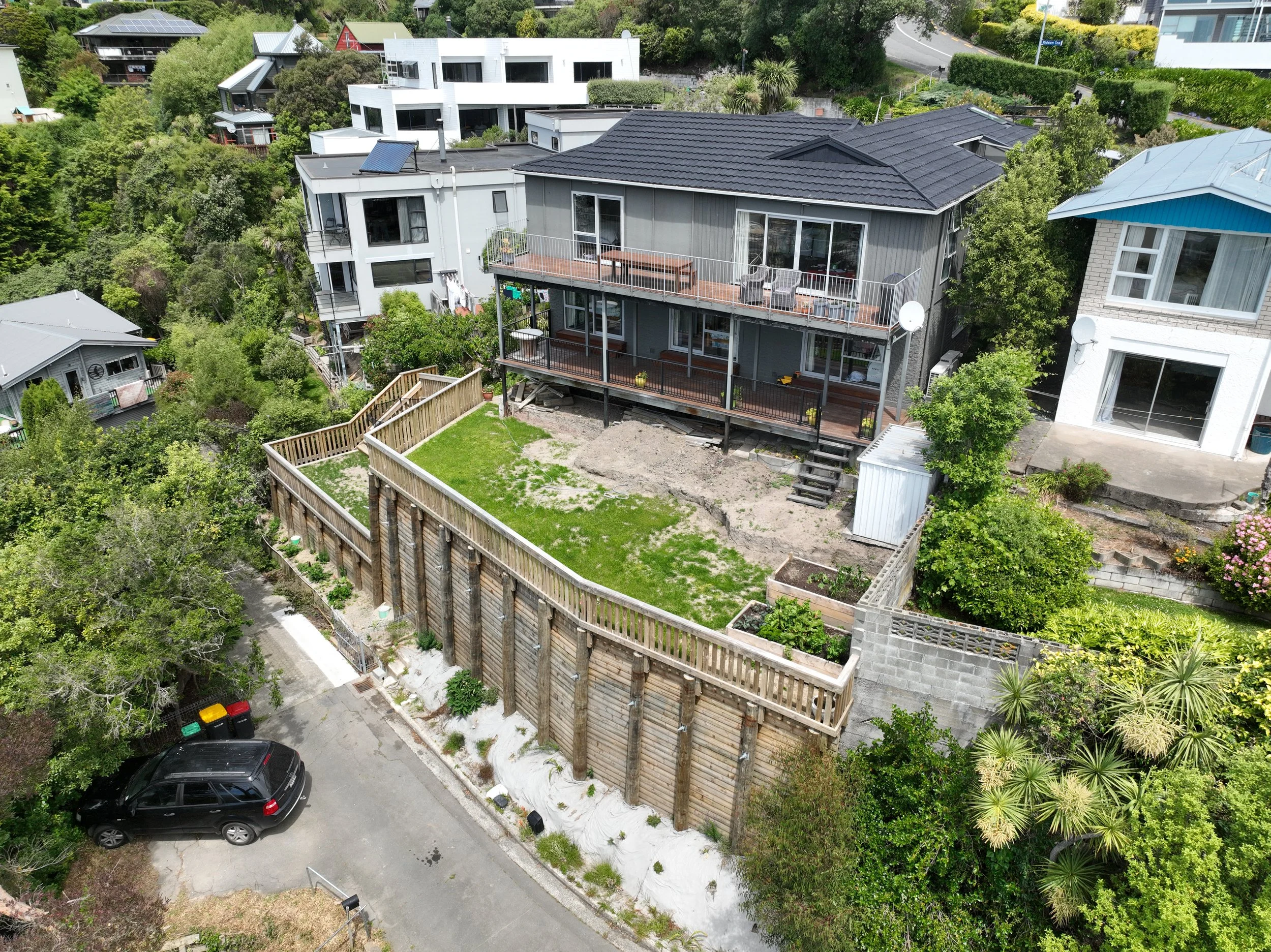 Aerial view of a residential neighborhood with modern houses, green trees, and a fenced backyard under construction, featuring a new deck being built on a gray house with a dark roof.